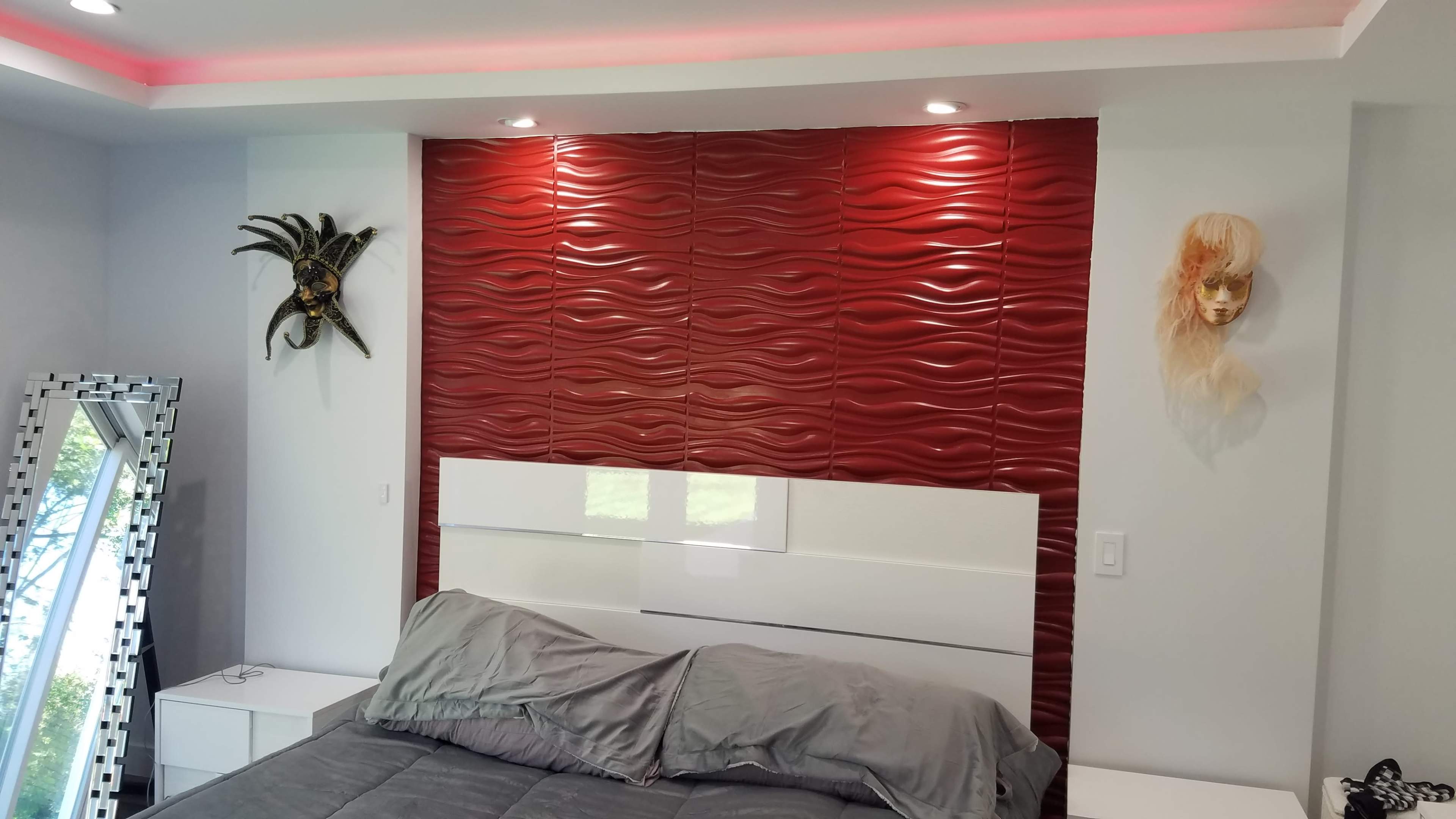 A modern bedroom with a red wavy textured accent wall behind a white bed, flanked by a decorative mask and a stylized face sculpture.