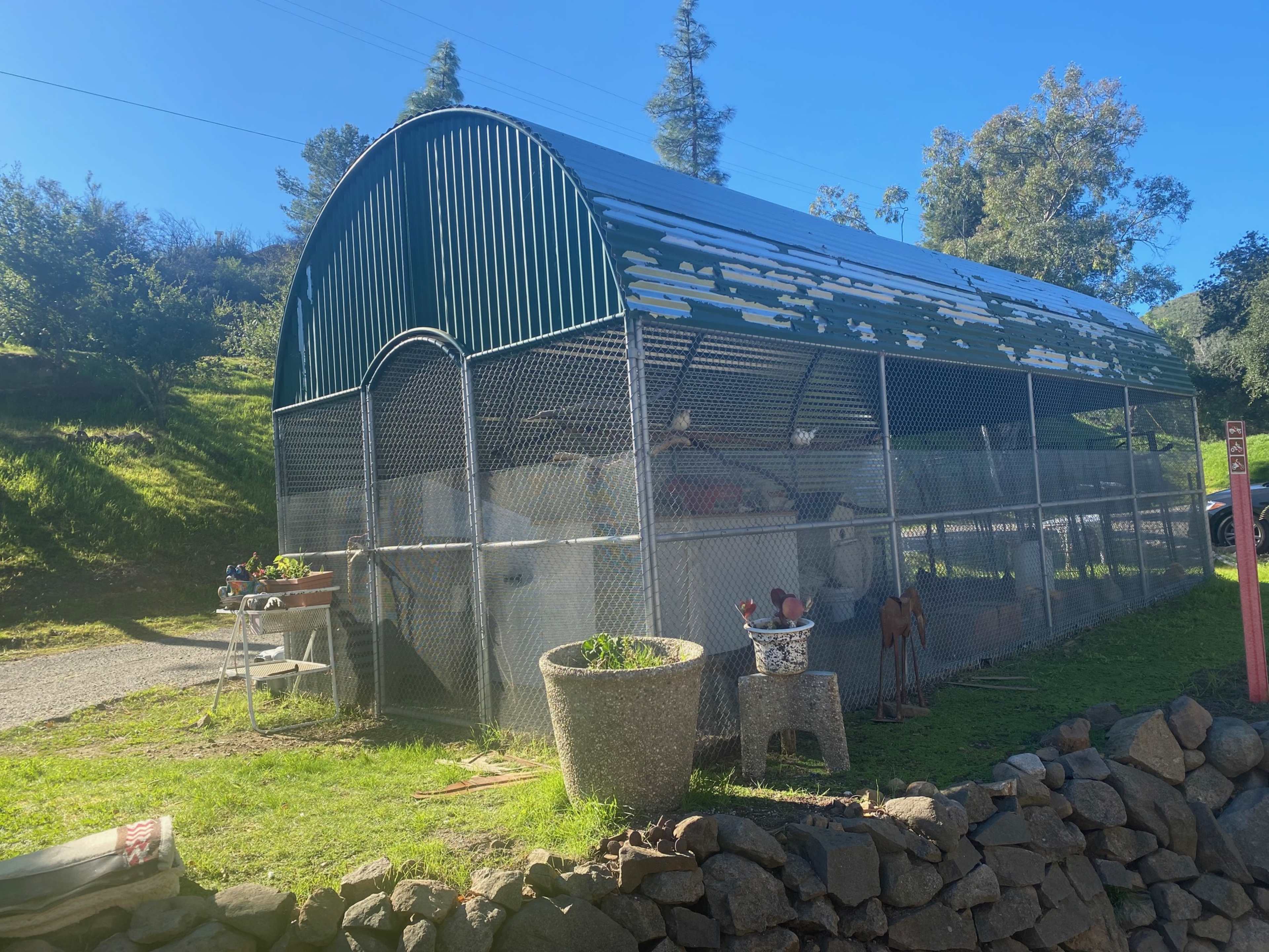 The image shows a large metal barn-like structure enclosed by a chain-link fence, situated on a grassy area beside a stone wall.
