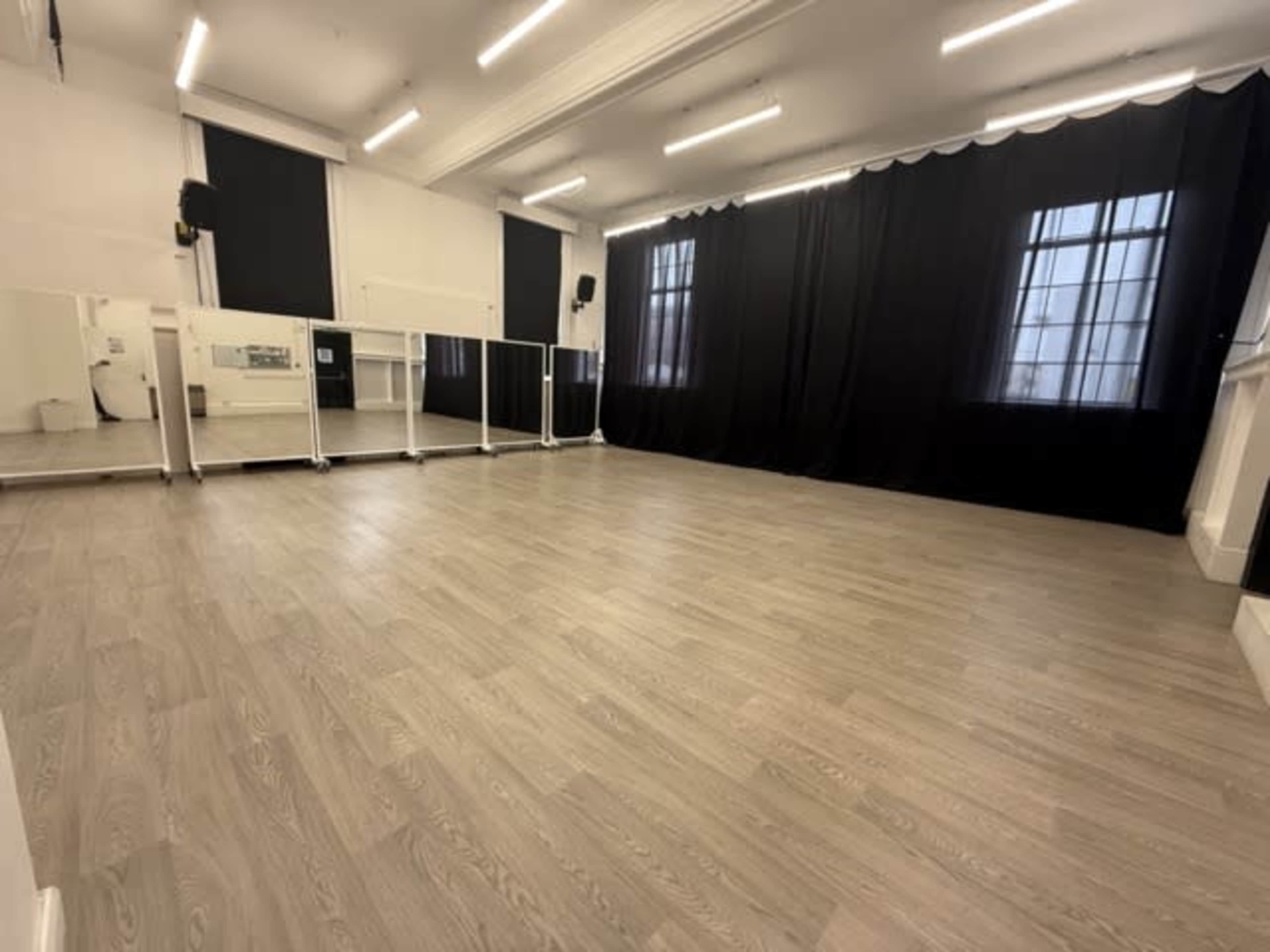 The image shows a spacious dance studio with wooden flooring, large mirrors on one wall, and black curtains at the windows.