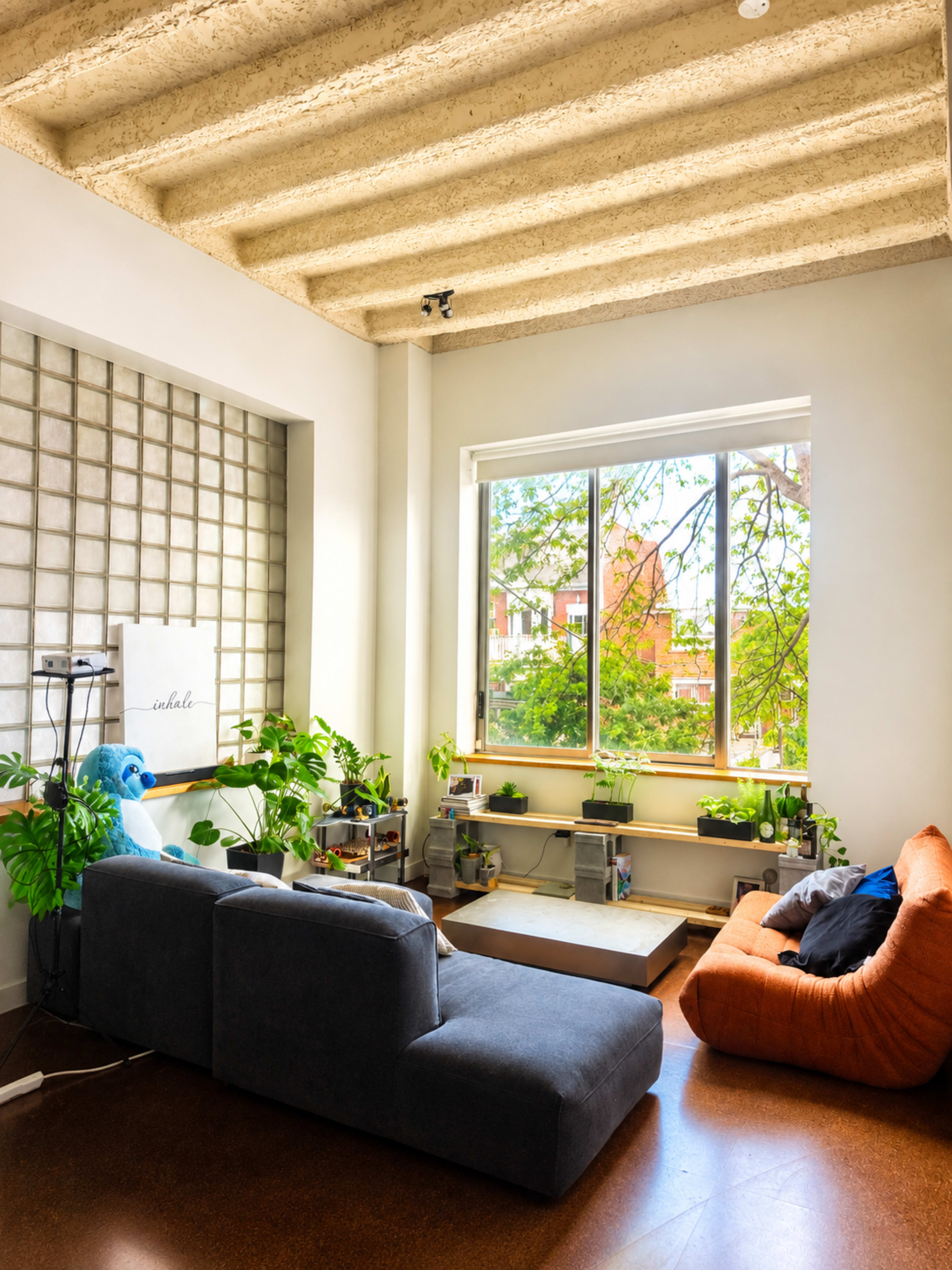 The image depicts a modern living room featuring a sectional sofa, potted plants, a large window with natural light, and concrete ceiling beams.