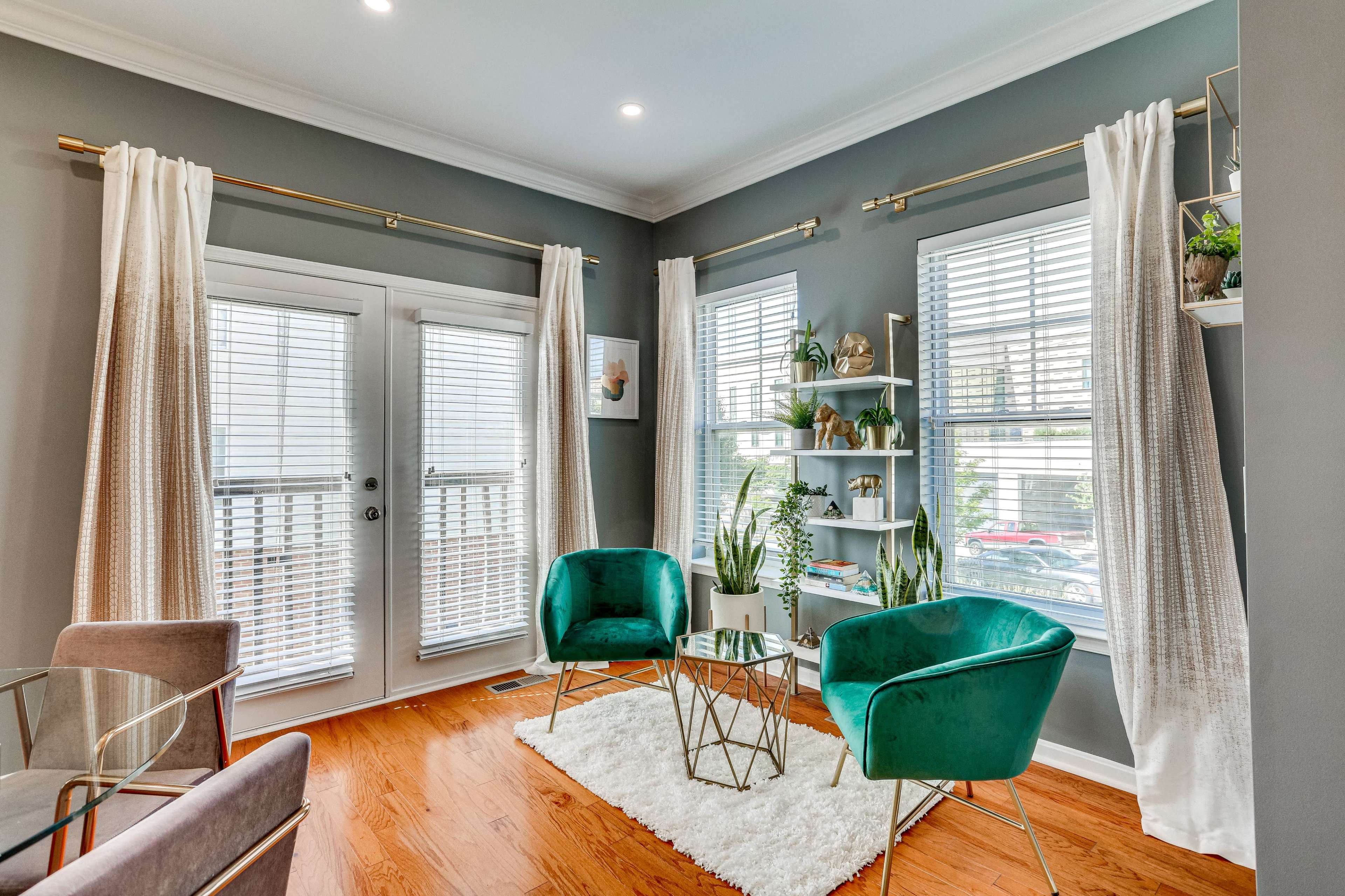 A well-lit corner of a stylish living room featuring two green chairs, a small gold coffee table, and decorative plants by the windows.