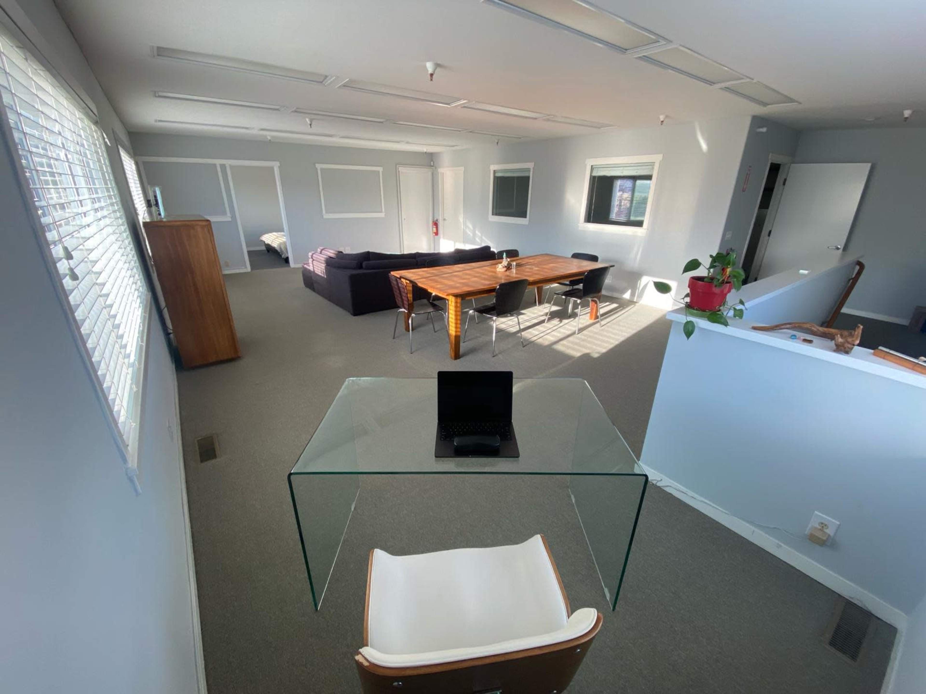 The image shows a spacious office or common area with a large wooden table, a black couch, multiple windows, and a glass desk with a tablet on it.