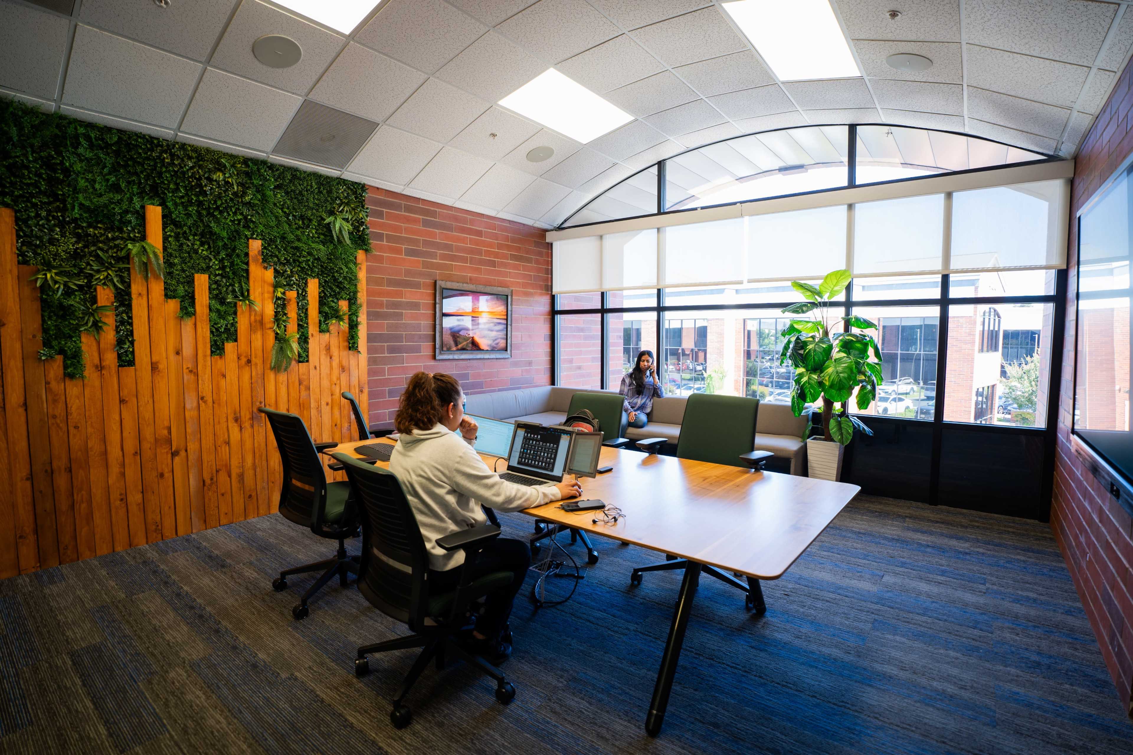 A modern office space features a woman working at a table while another woman sits nearby by a large window with plants and wood paneling.