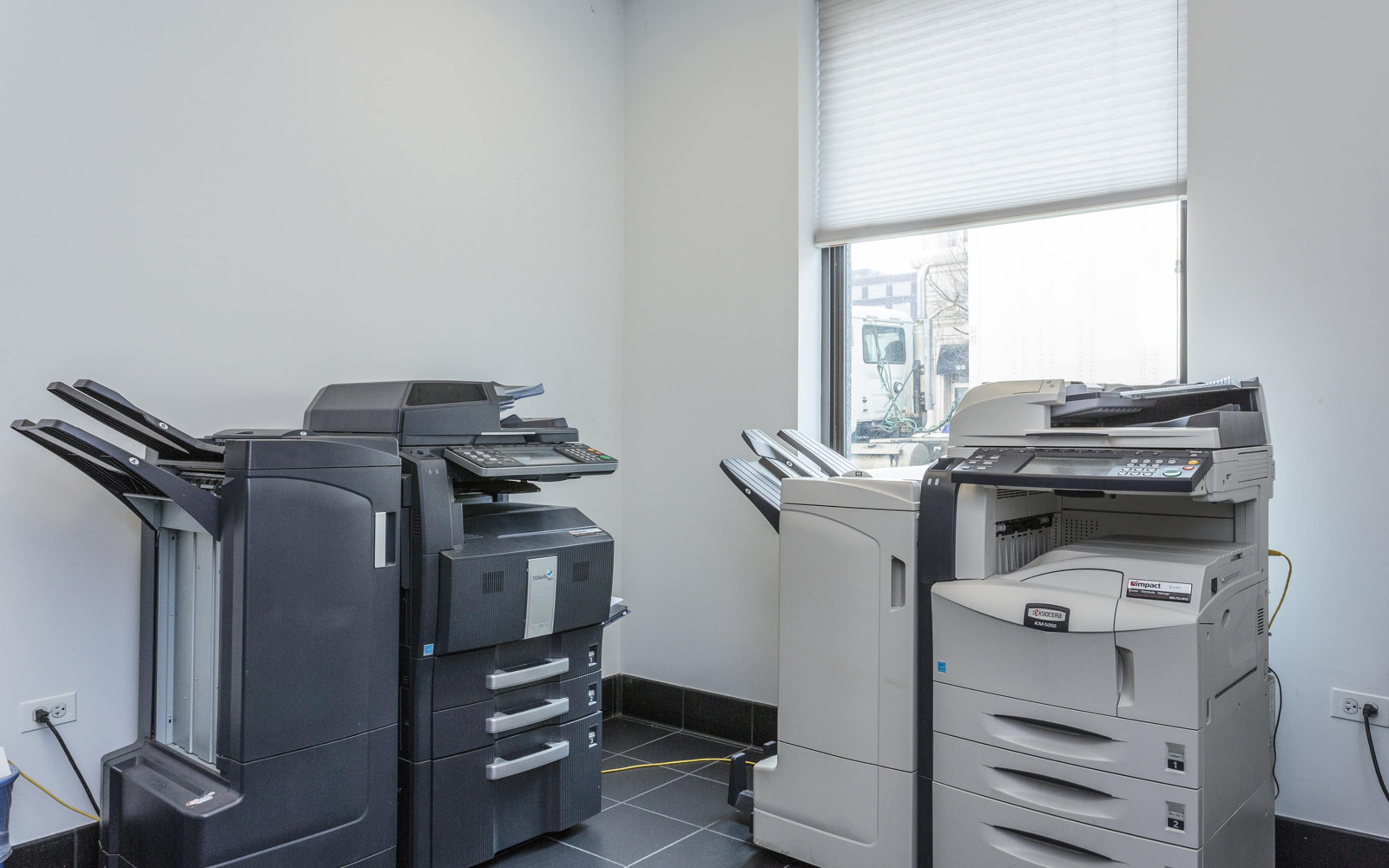 A small office space with several photocopiers and printers positioned against a wall under a window.