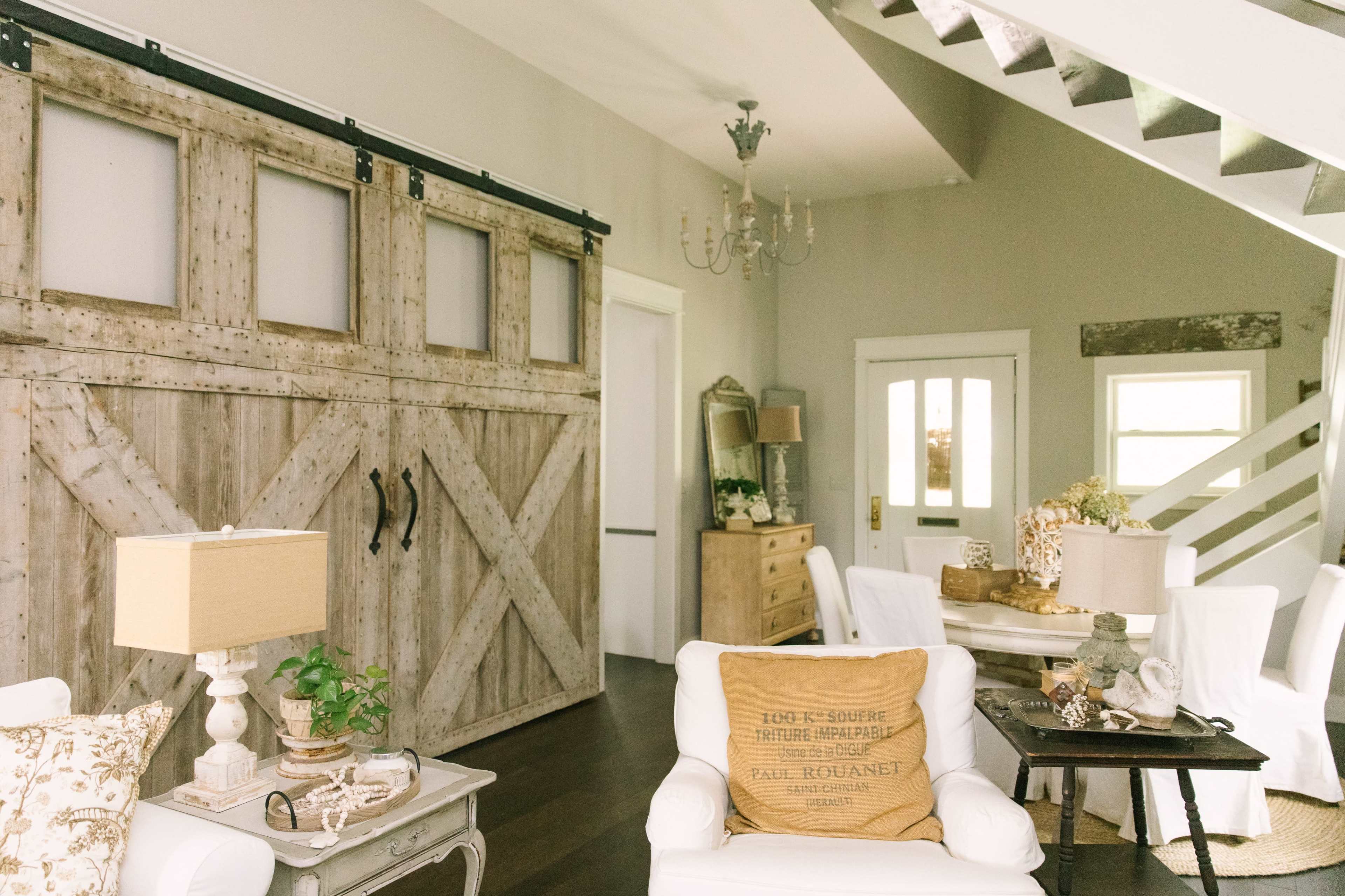 The image shows a light-filled living space featuring a rustic barn door, a dining area with a small table, and white furniture arranged around a wooden accent table.