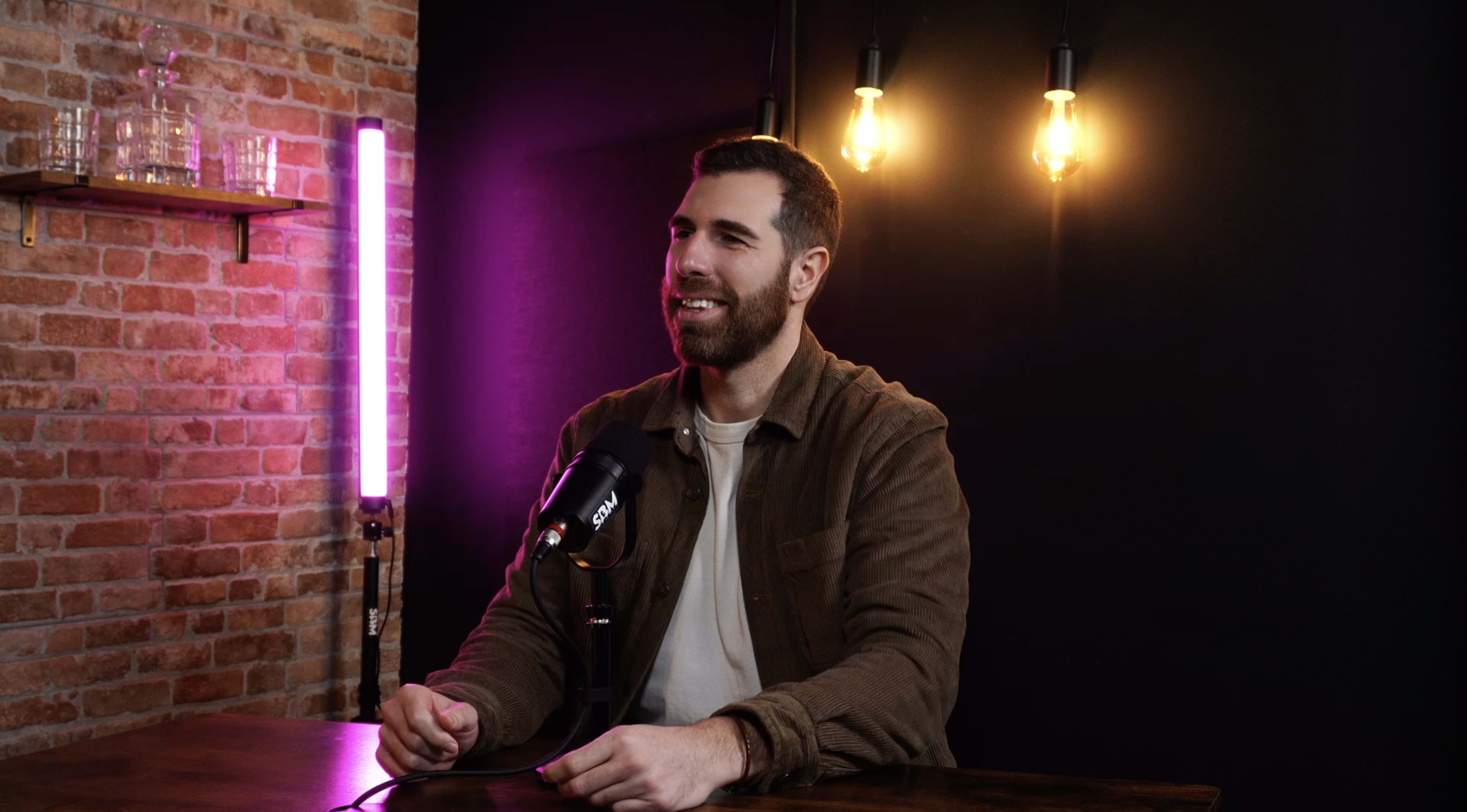 A man sits at a table with a microphone in front of him, smiling against a backdrop of brick and illuminated lights.