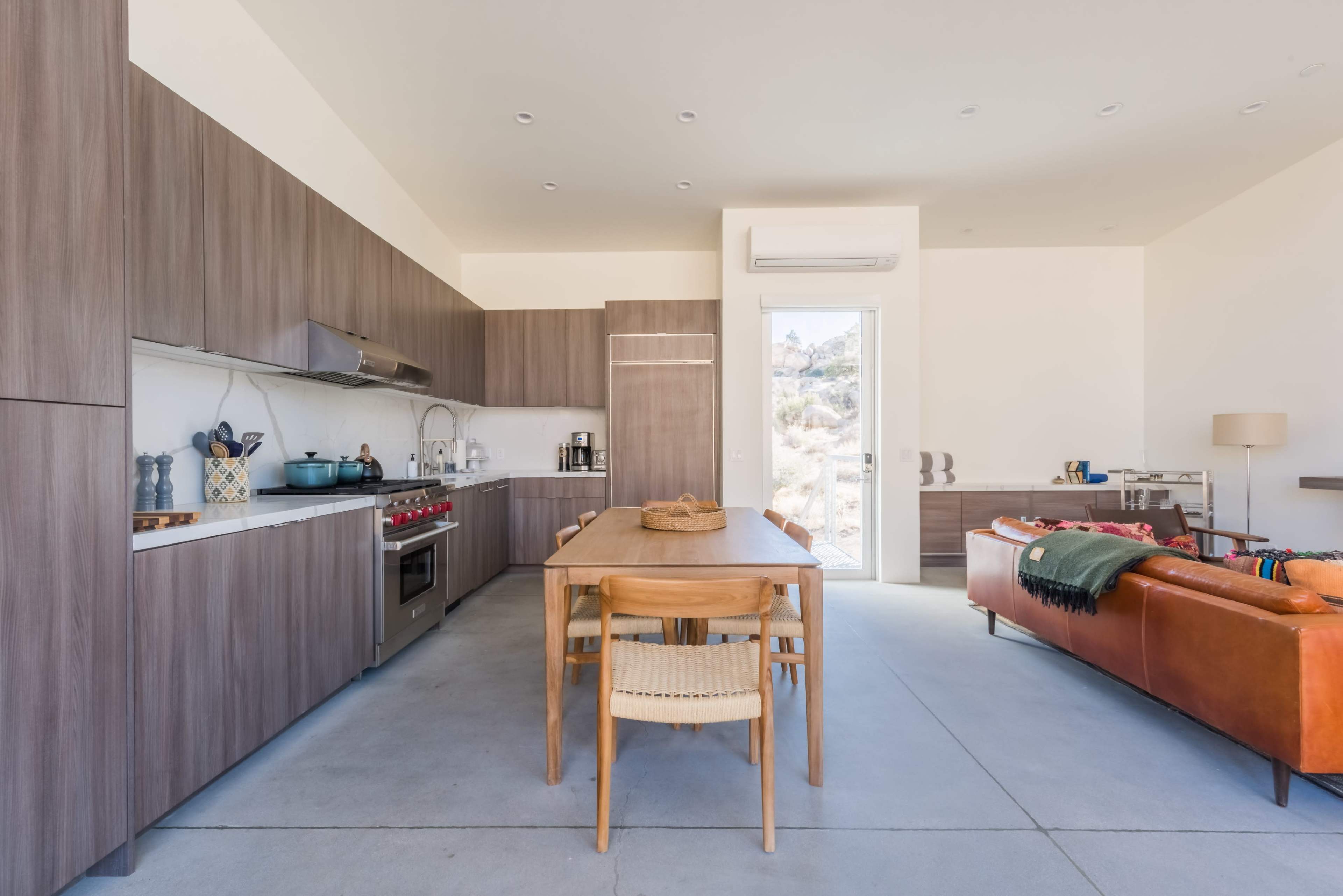 The image shows a modern kitchen and dining area featuring a long wooden table surrounded by chairs, a sleek kitchen with a gas stove, and a cozy living space with an orange couch.