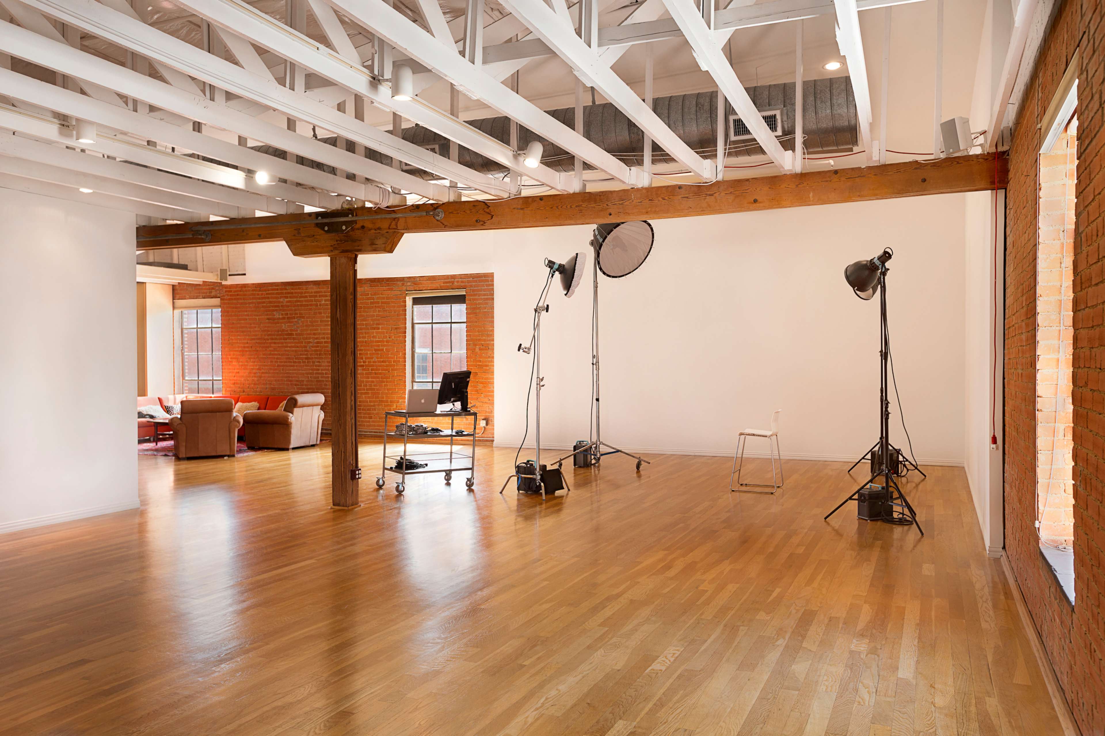 An open space with hardwood floors, exposed wooden beams, and studio lighting equipment set up on one side, while a cozy seating area is visible in the background.