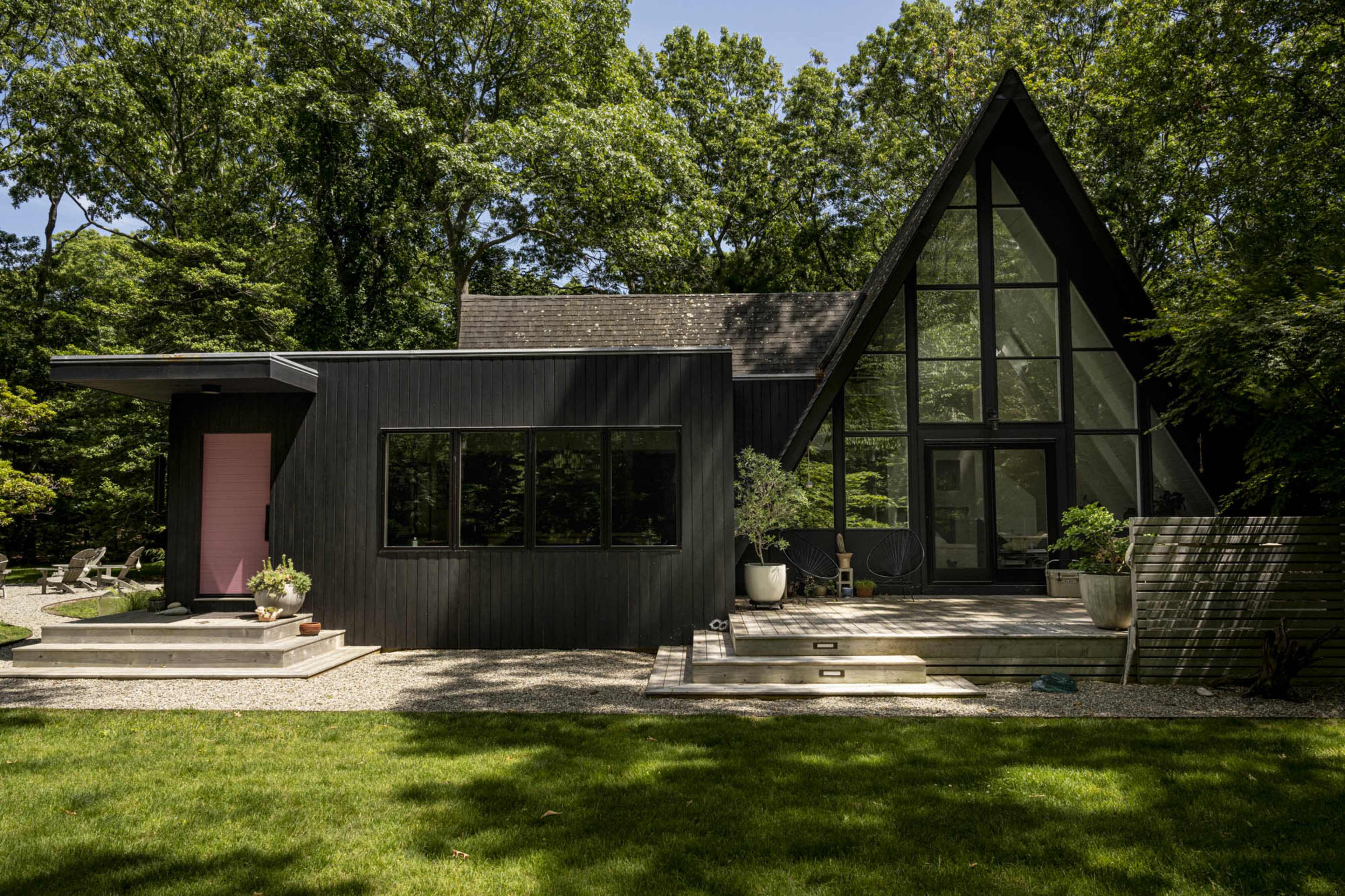 The image shows a modern black wooden house with a large glass facade, surrounded by greenery and featuring a stone pathway and patio area.