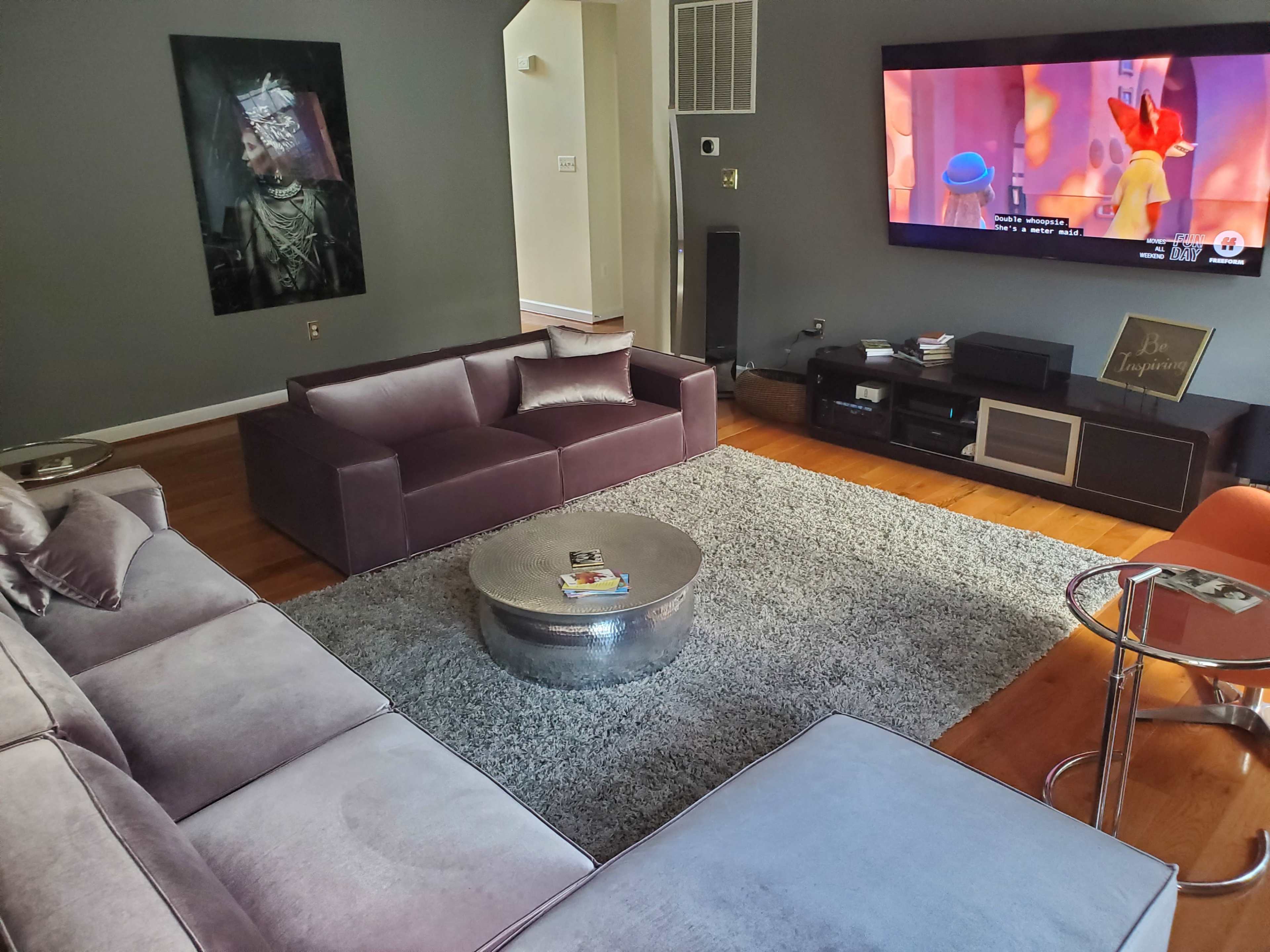 A modern living room with a grey sectional sofa, a round metallic coffee table, a large television displaying a colorful scene, and a plush rug on wooden flooring.