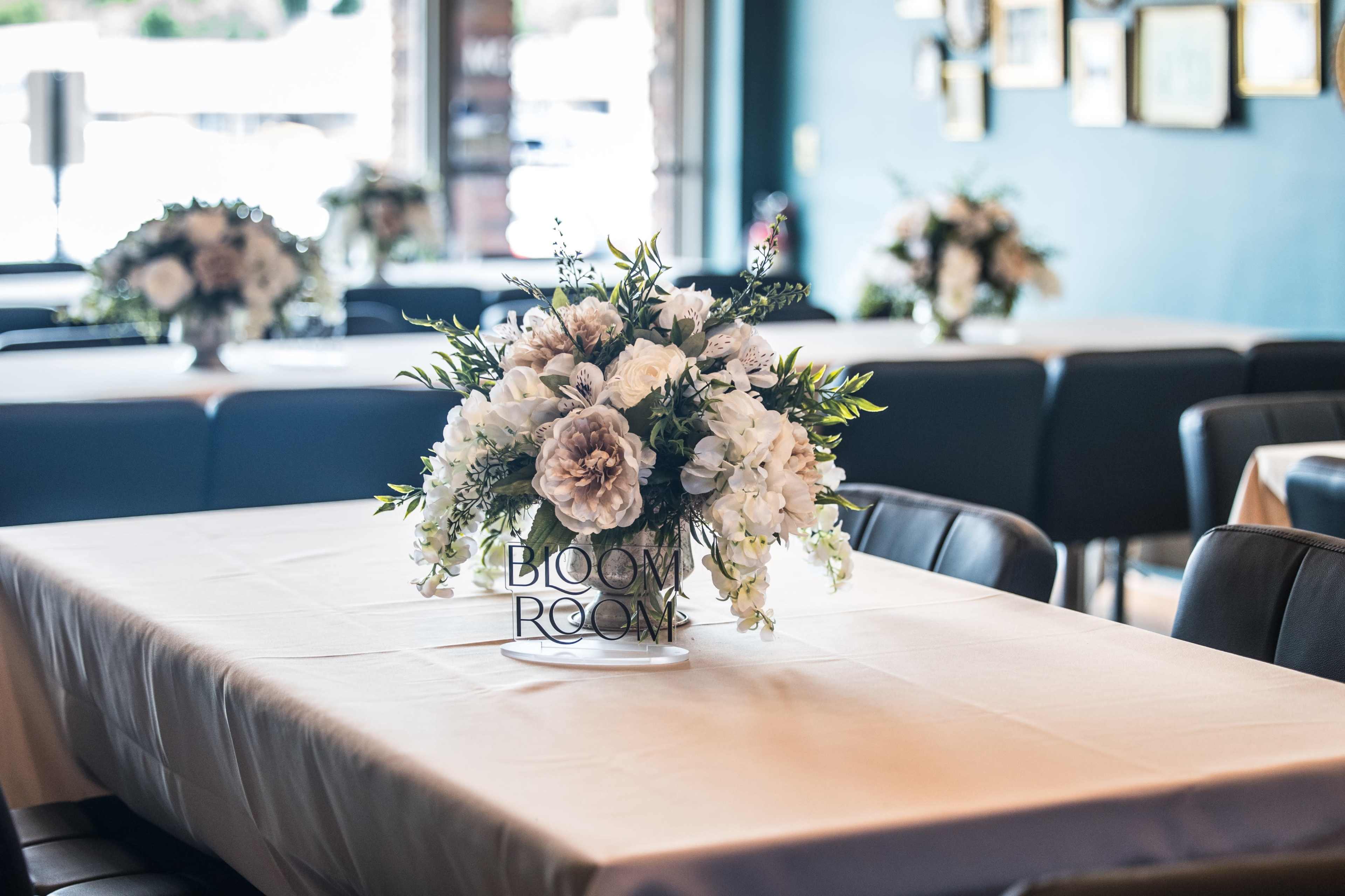 A table in a dining area is set with a floral centerpiece and a sign that reads "BLOOM ROOM."