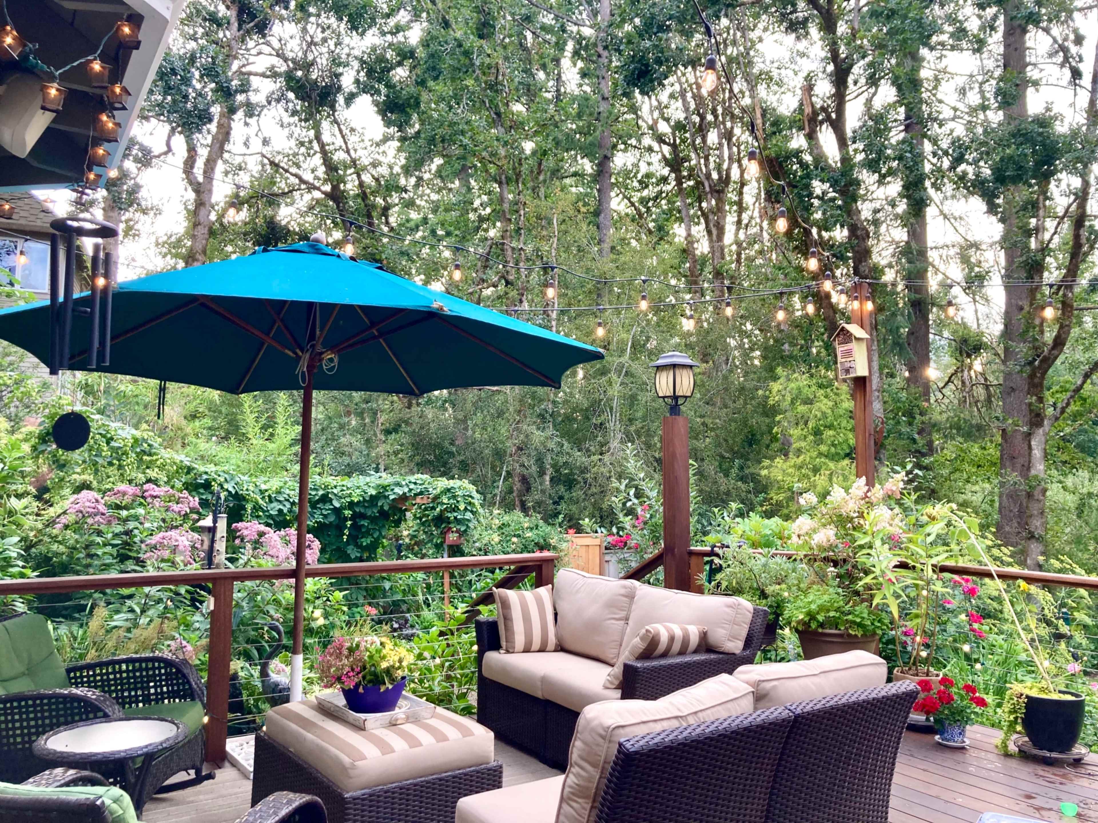 A patio area is furnished with a seating arrangement under a large umbrella, surrounded by greenery and string lights.