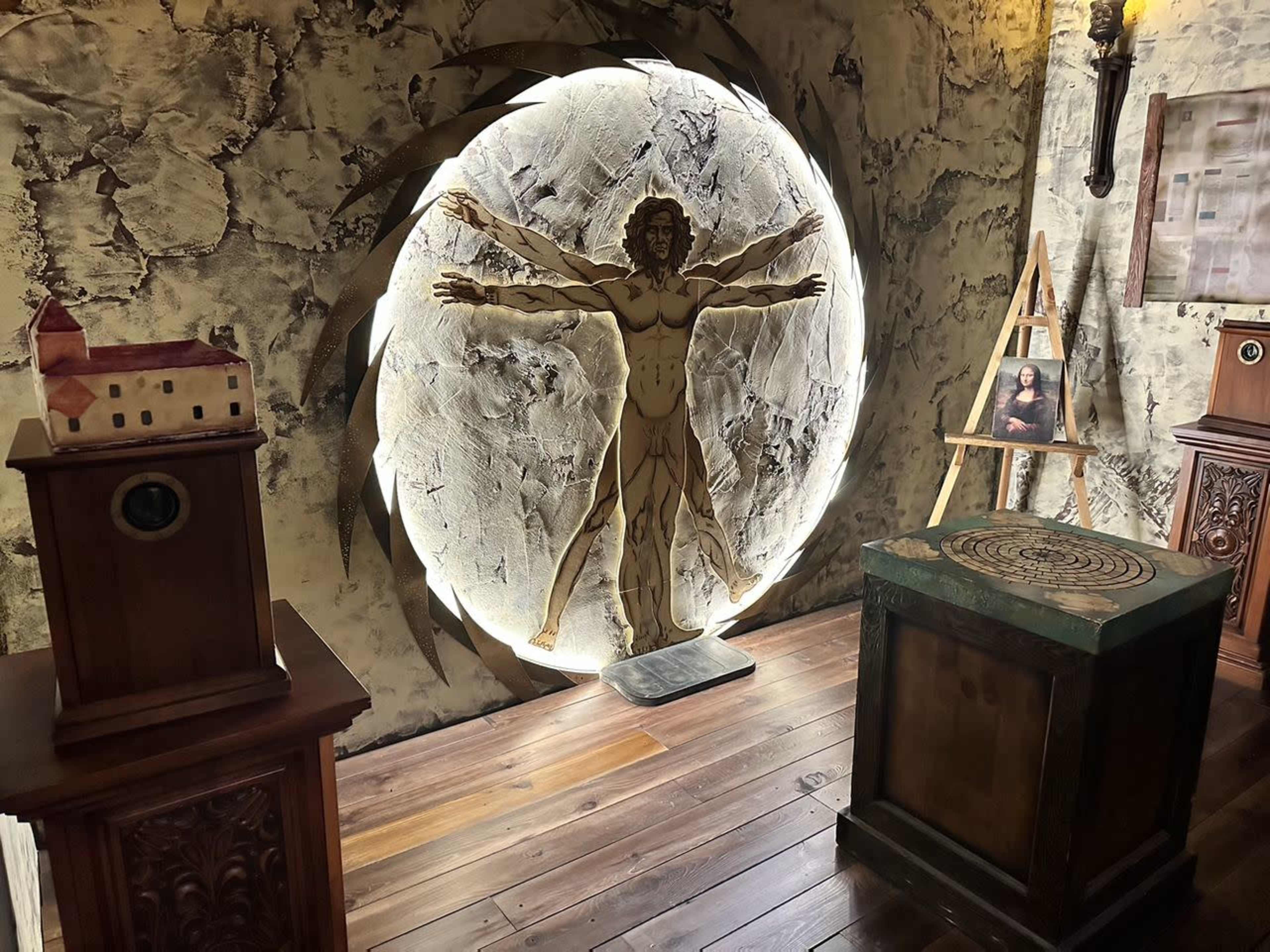 The image features a room with a circular illuminated display of a human figure inspired by Leonardo da Vinci's Vitruvian Man, surrounded by wooden furniture and artwork.