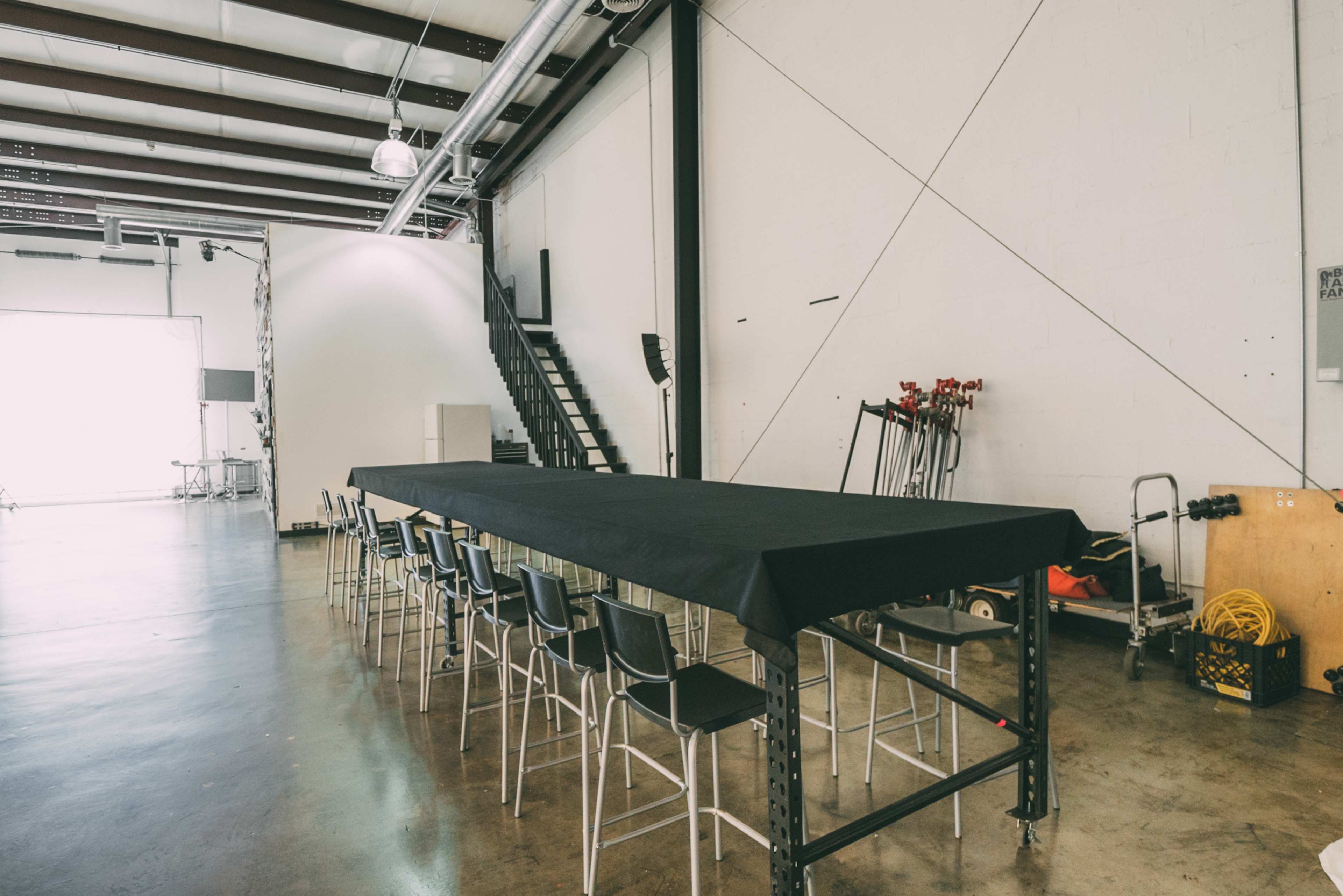 A long black table with chairs is set up in an industrial-style room with a staircase and equipment in the background.