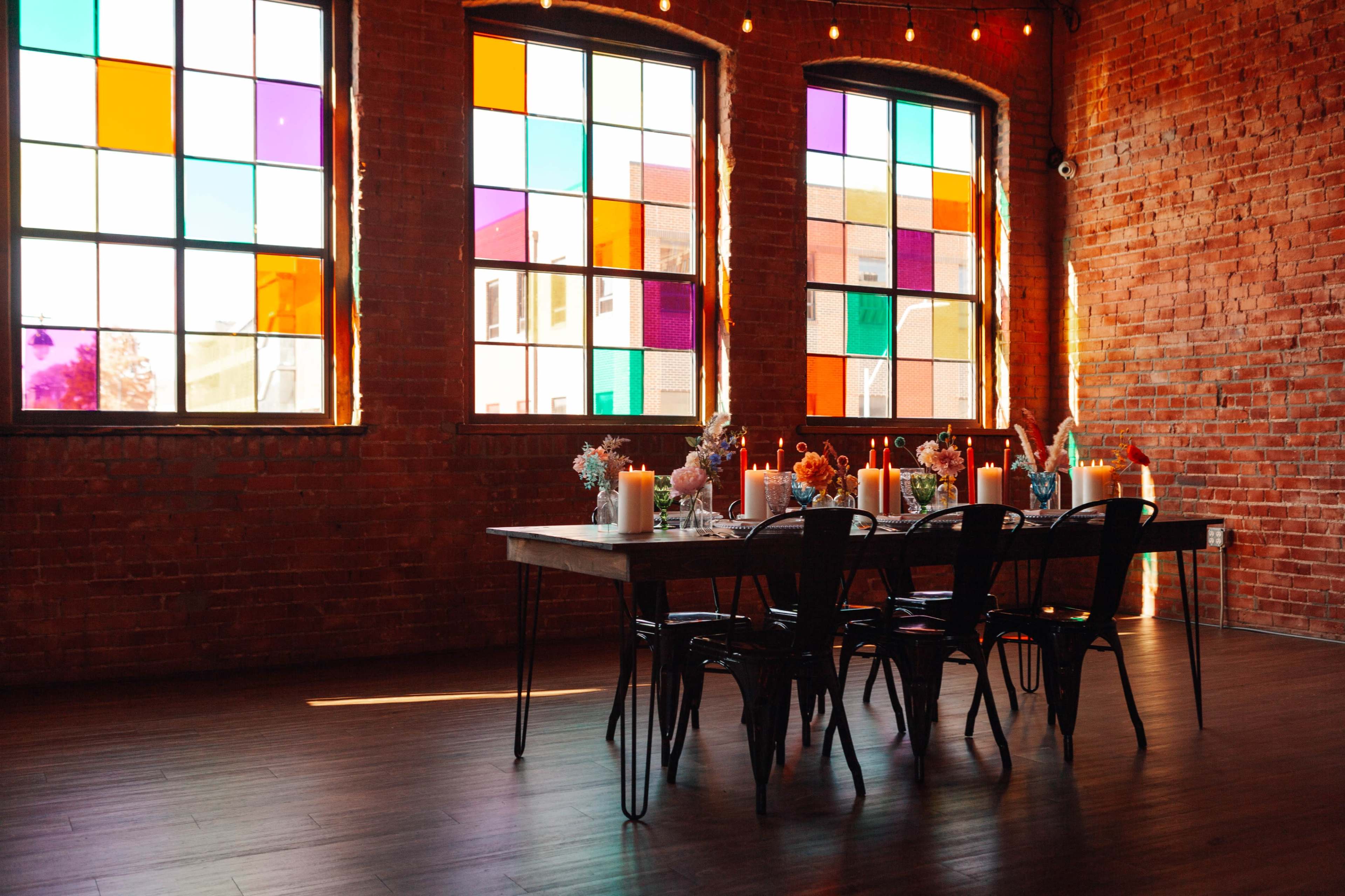 A long dining table is set with candles and floral arrangements in a brick-walled room featuring large, colorful windows.