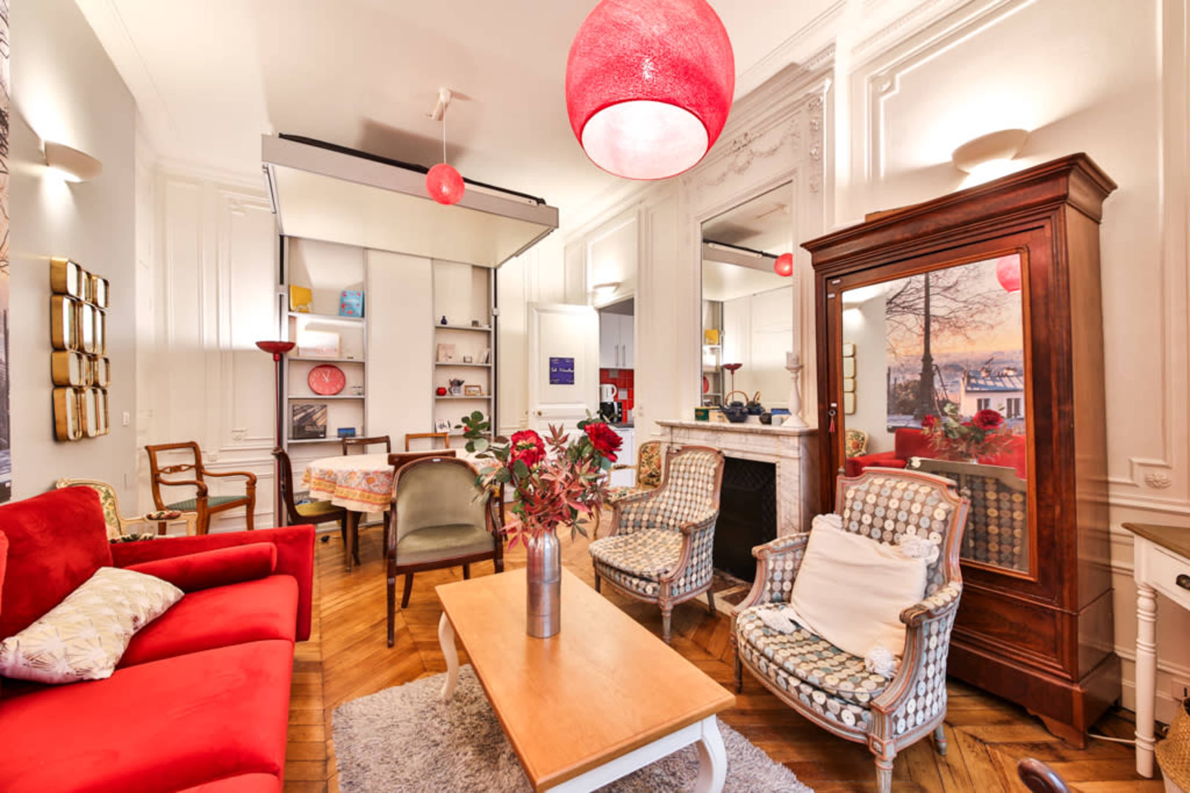 The image depicts a stylish living room featuring a red couch, two patterned armchairs, a wooden coffee table, and a fireplace surrounded by decorative items.