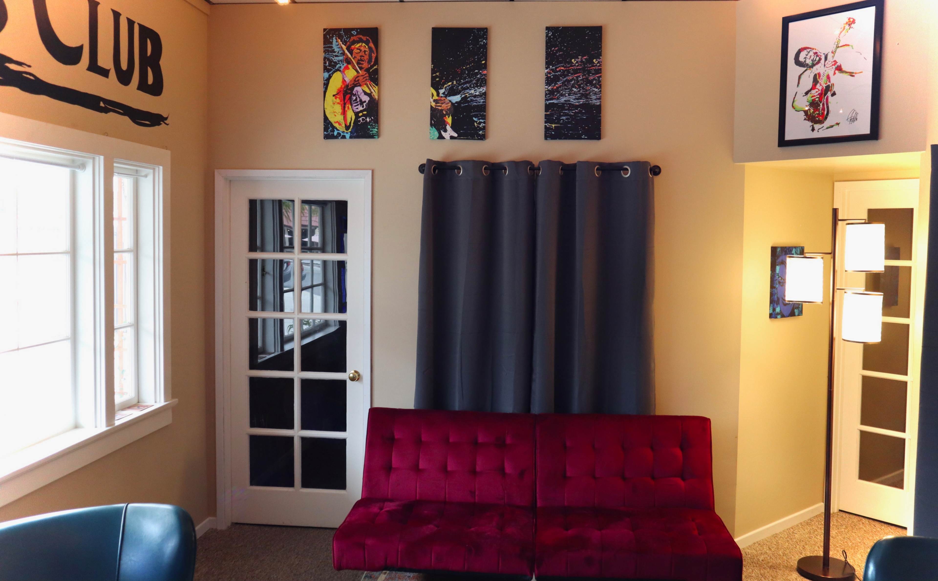 The image shows a cozy interior with a red tufted couch, gray curtains, framed artwork on the walls, and a white door leading to another room.