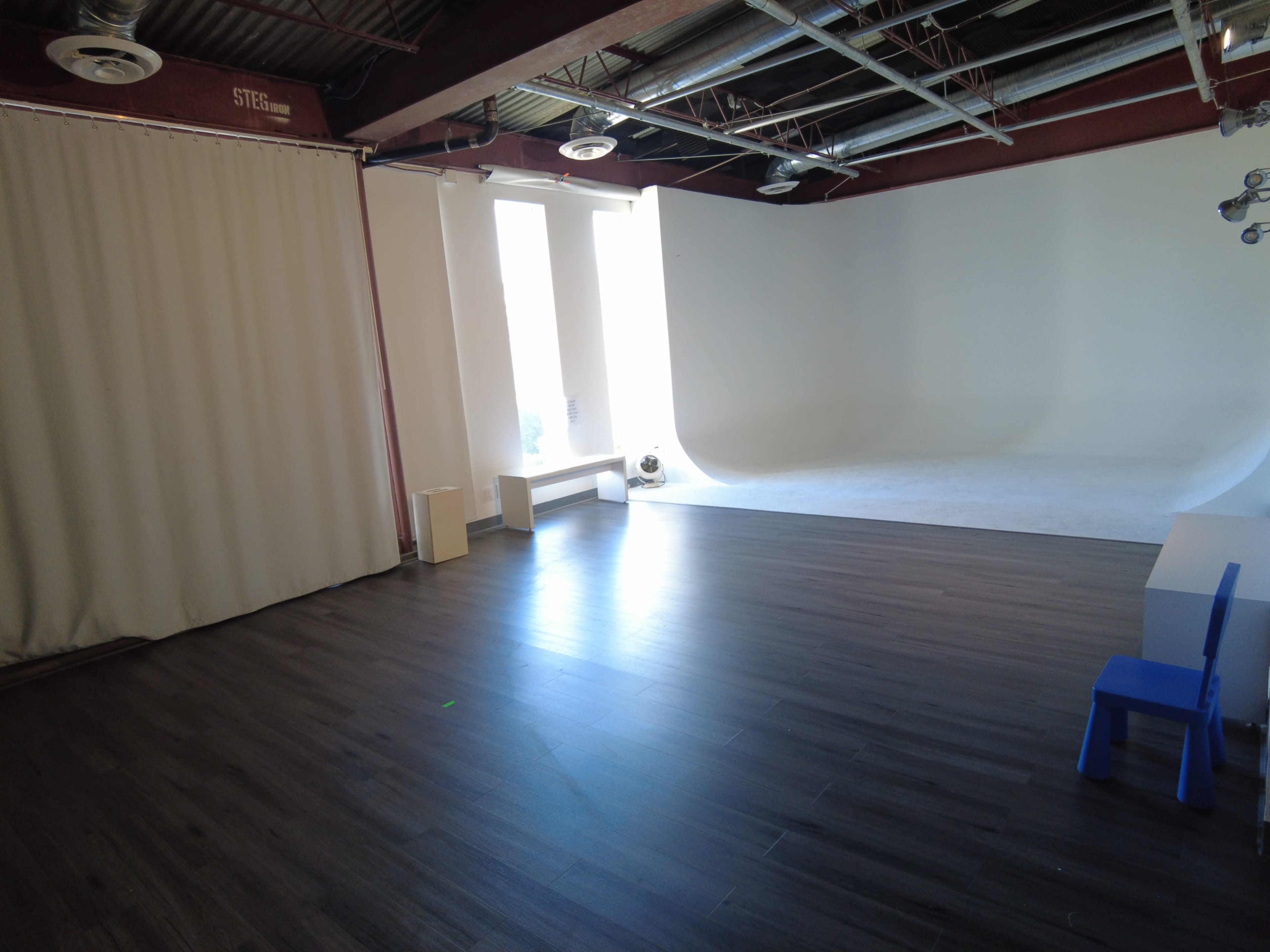 The image shows a spacious photography studio with a hardwood floor, large windows allowing natural light, a blank backdrop, and minimal furniture including a small blue chair.