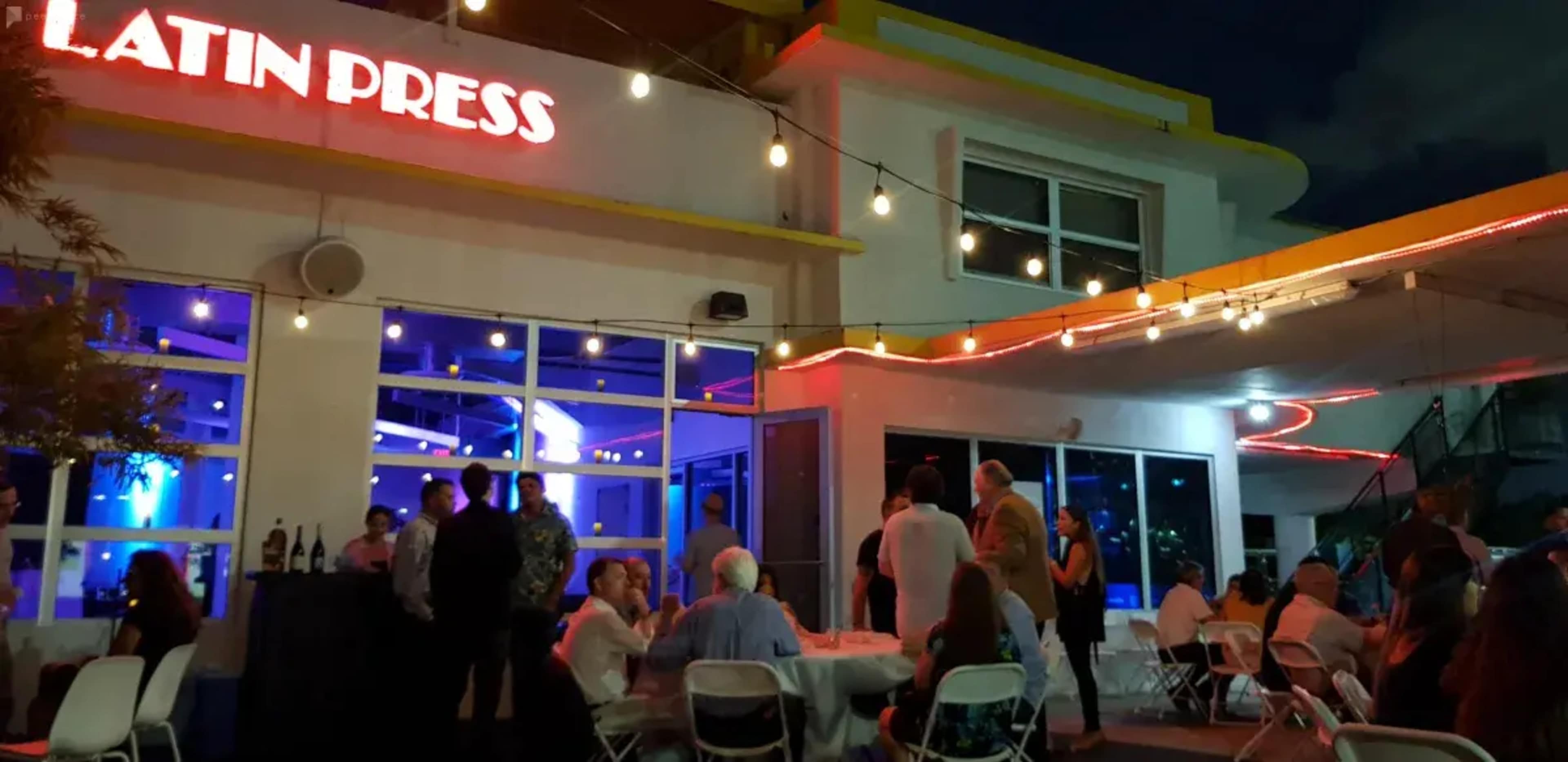 A gathering takes place outside a building labeled "Latin Press," with attendees seated at tables and mingling under string lights.