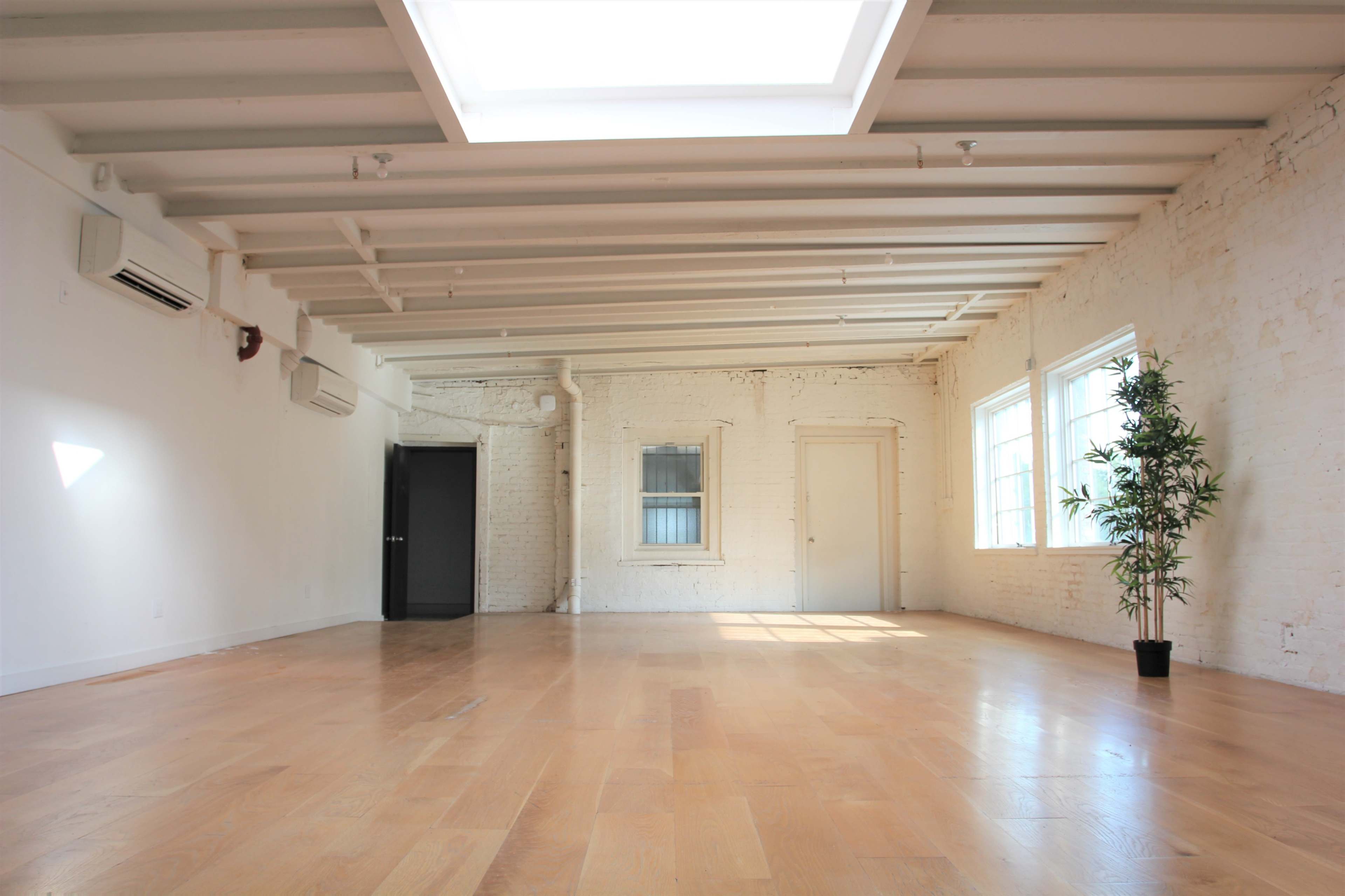 The image shows a spacious, empty room with wooden flooring, white brick walls, and a skylight overhead.