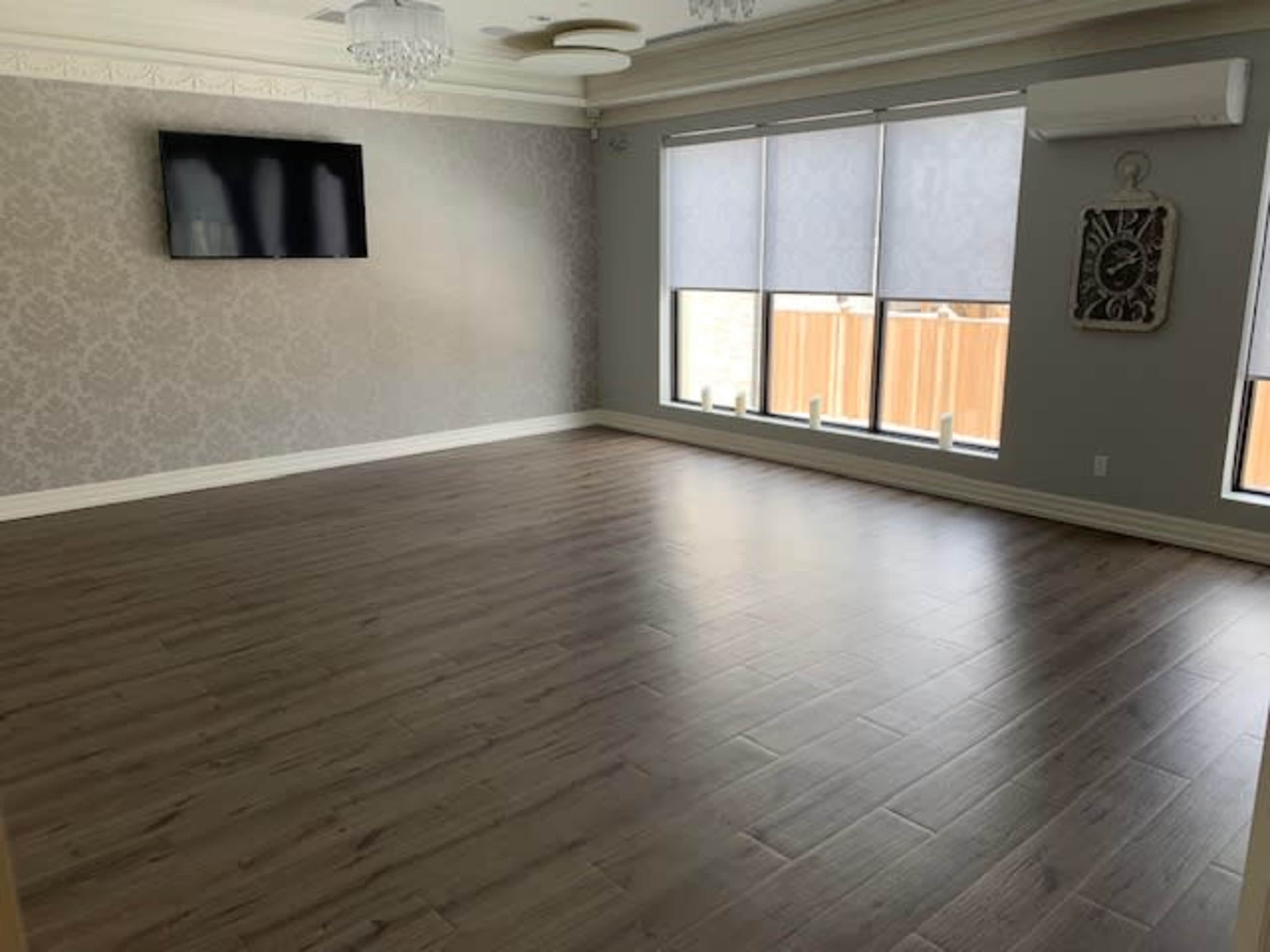 A spacious room with wooden flooring, a television mounted on the wall, and large windows letting in light.