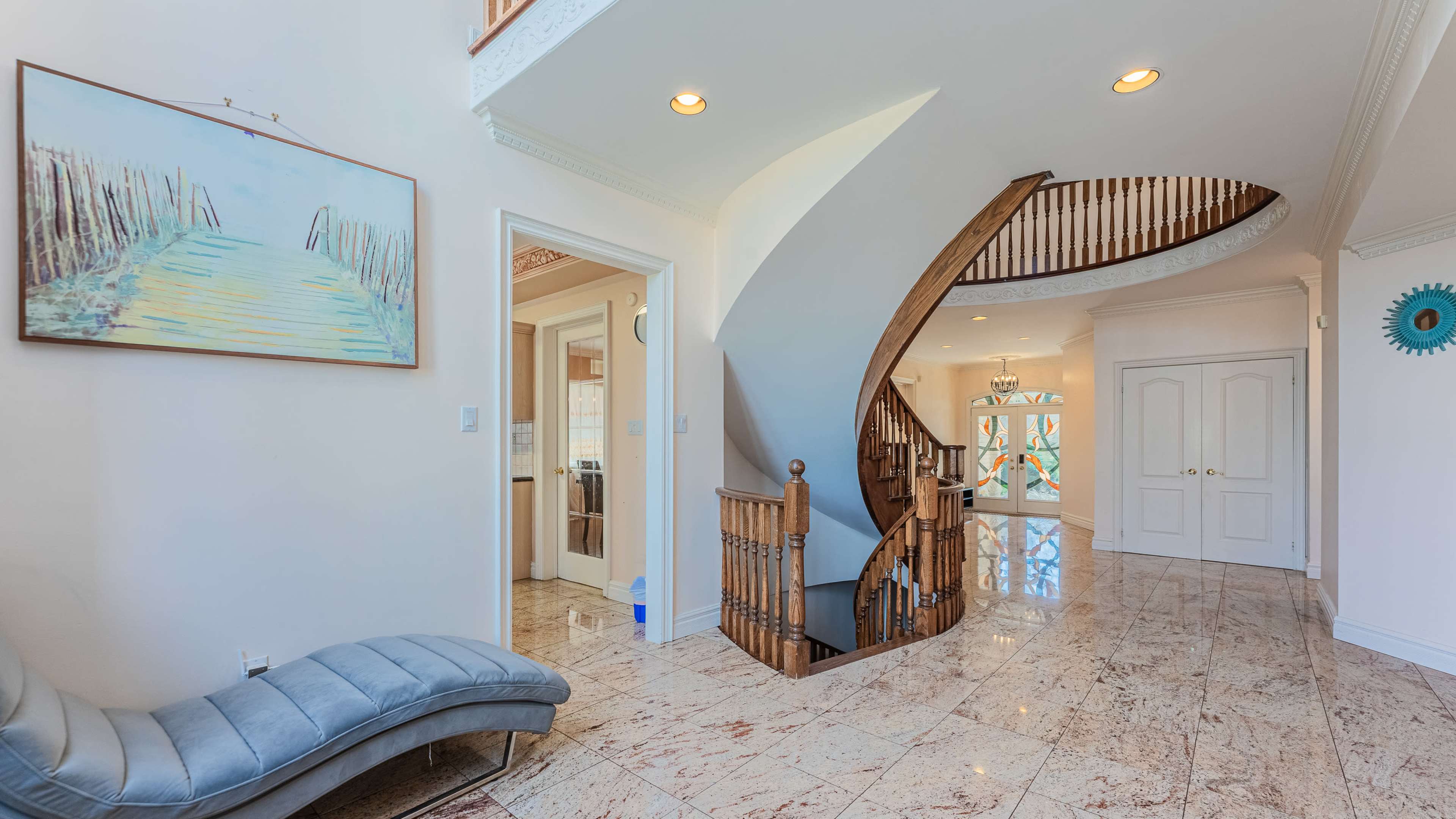 The image shows a foyer with a curved staircase and a piece of wall art depicting a pathway, accompanied by marble flooring and a bright ambiance.