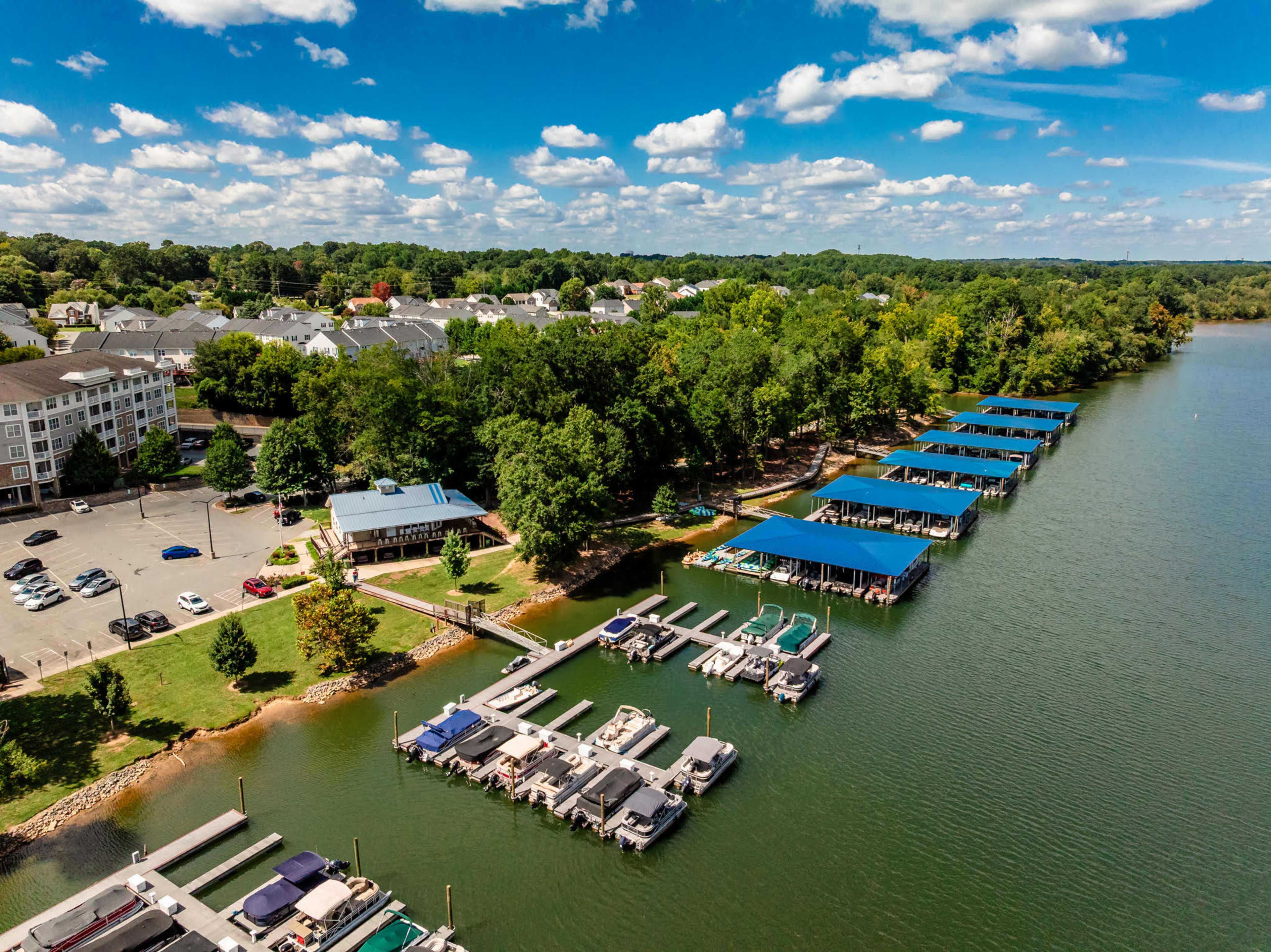 The image shows a marina with several boat docks alongside a green shoreline, a parking area, and residential buildings in the background under a partly cloudy sky.