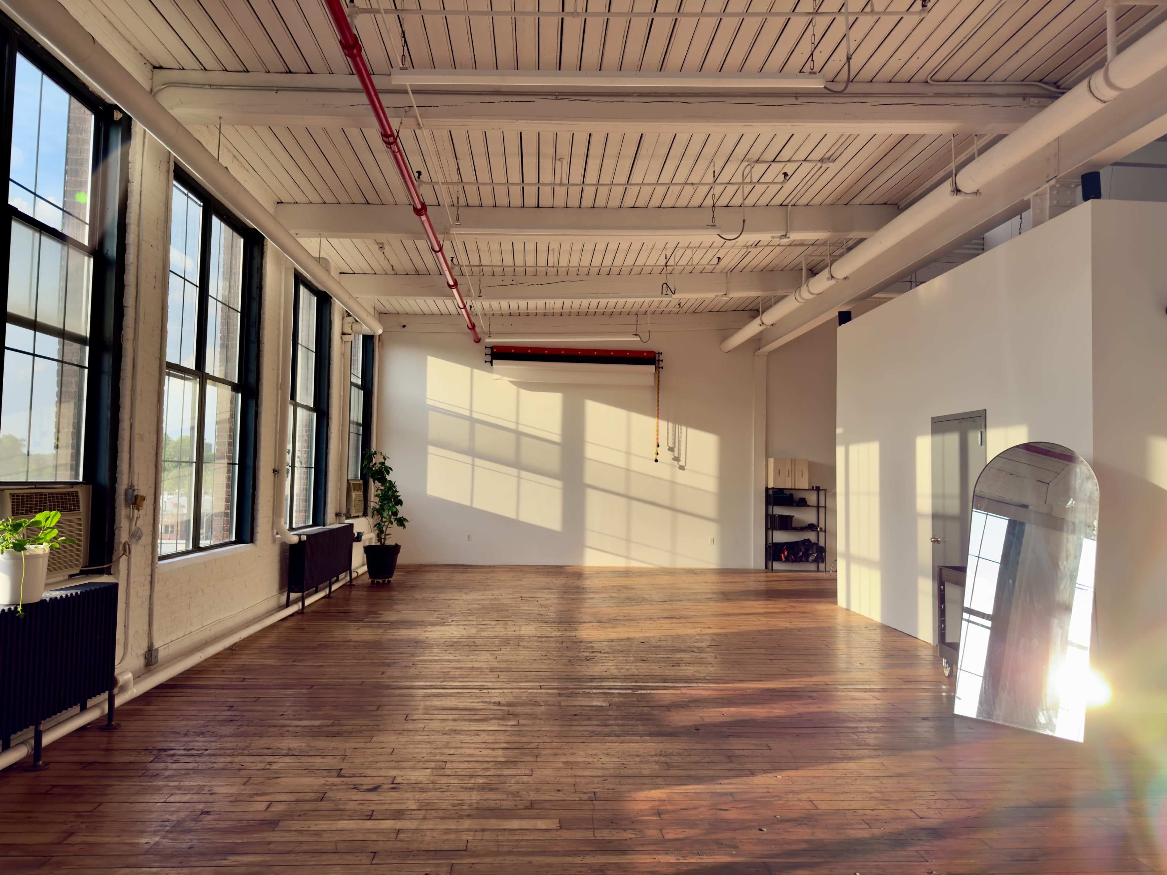 The image shows a spacious, well-lit room with large windows, wooden flooring, and sparse furnishings.