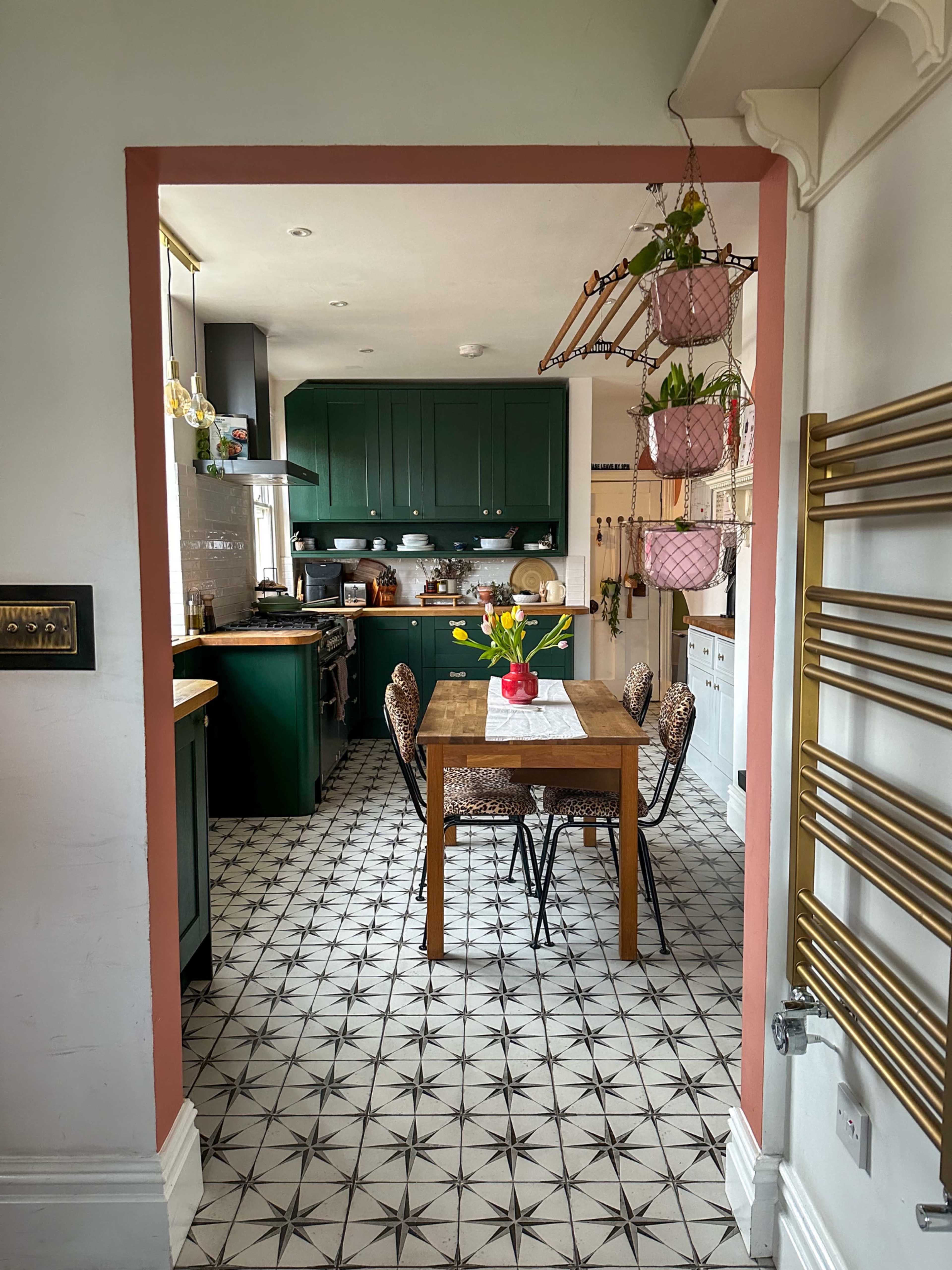 A kitchen with dark green cabinetry, a wooden dining table at the center, and patterned tile flooring is visible through an archway.