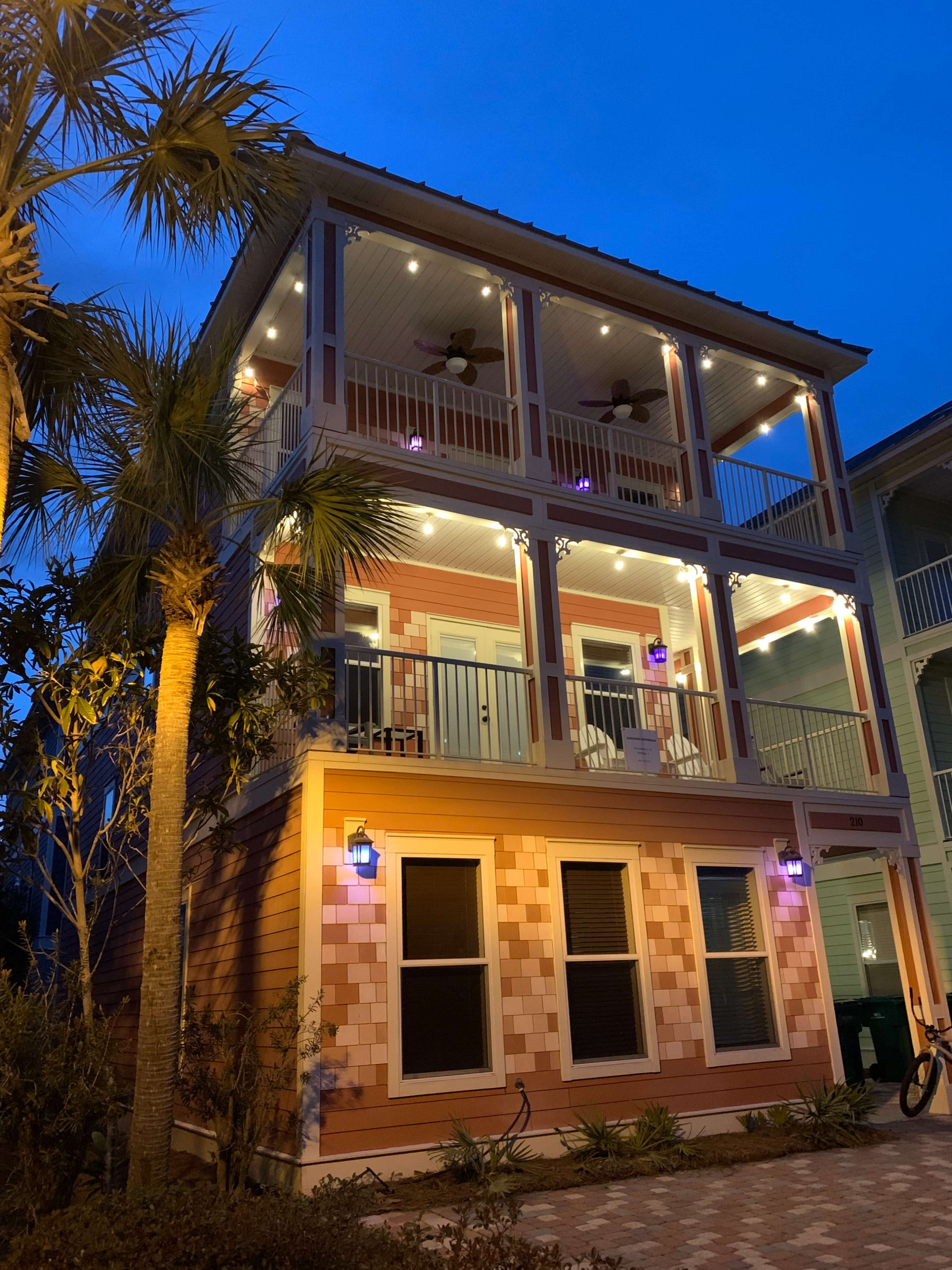 A three-story house with illuminated balconies and palm trees in the foreground is set against a twilight sky.