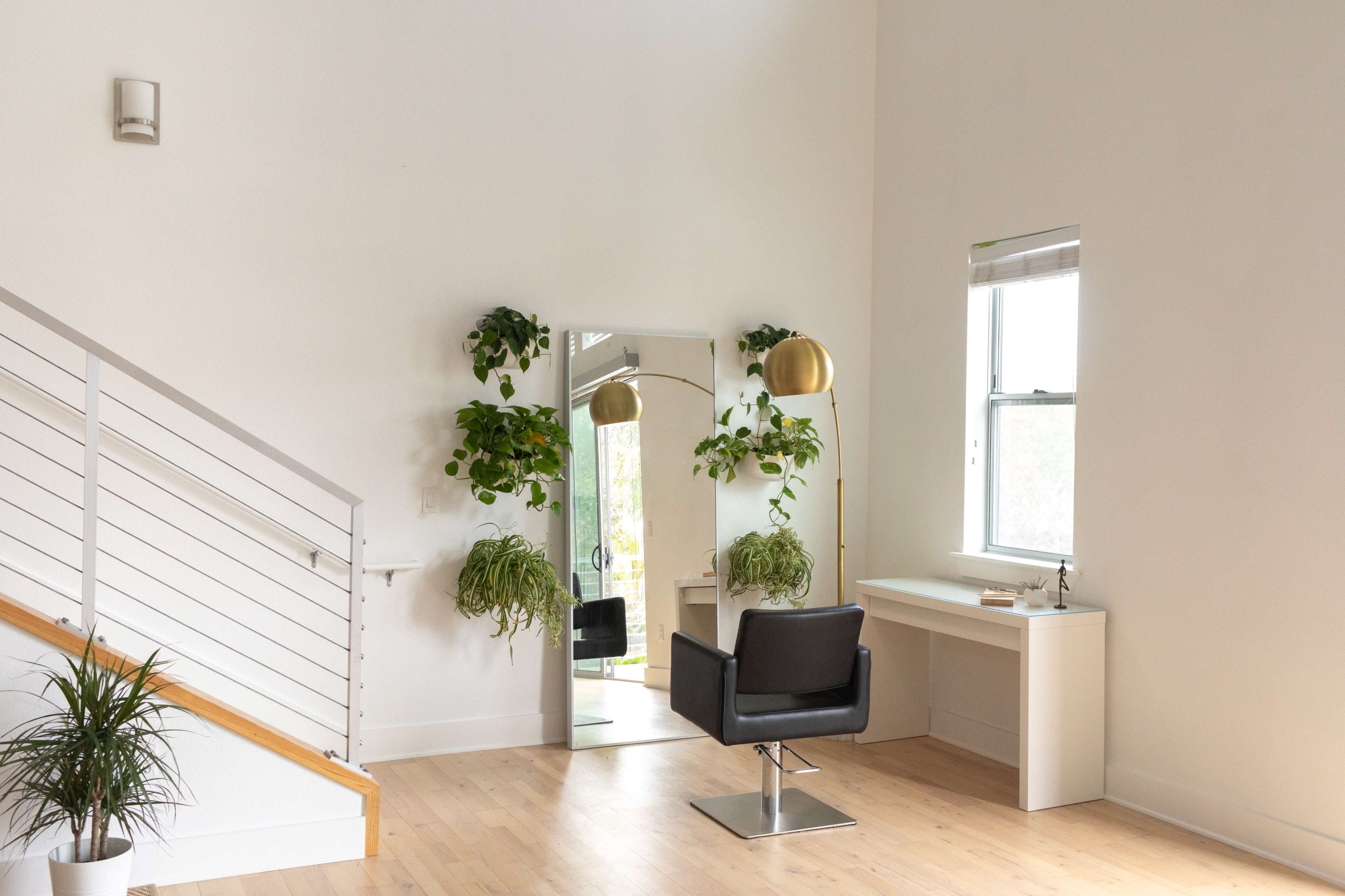 A minimalist room features a hair salon chair, a large mirror, and a desk beside windows adorned with green plants.