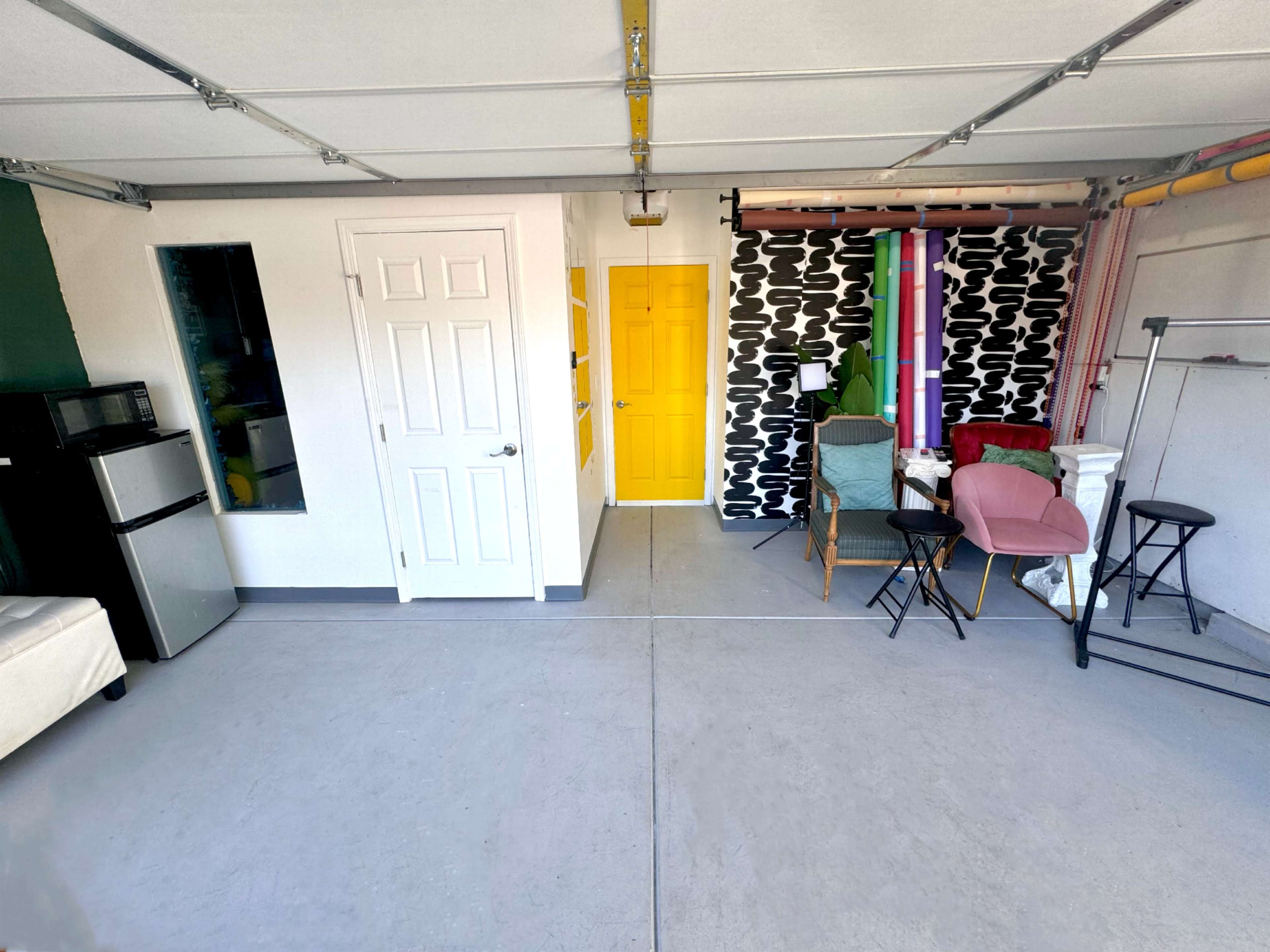 The image shows a spacious garage with a yellow door, a small refrigerator, and various seating options arranged along the walls.