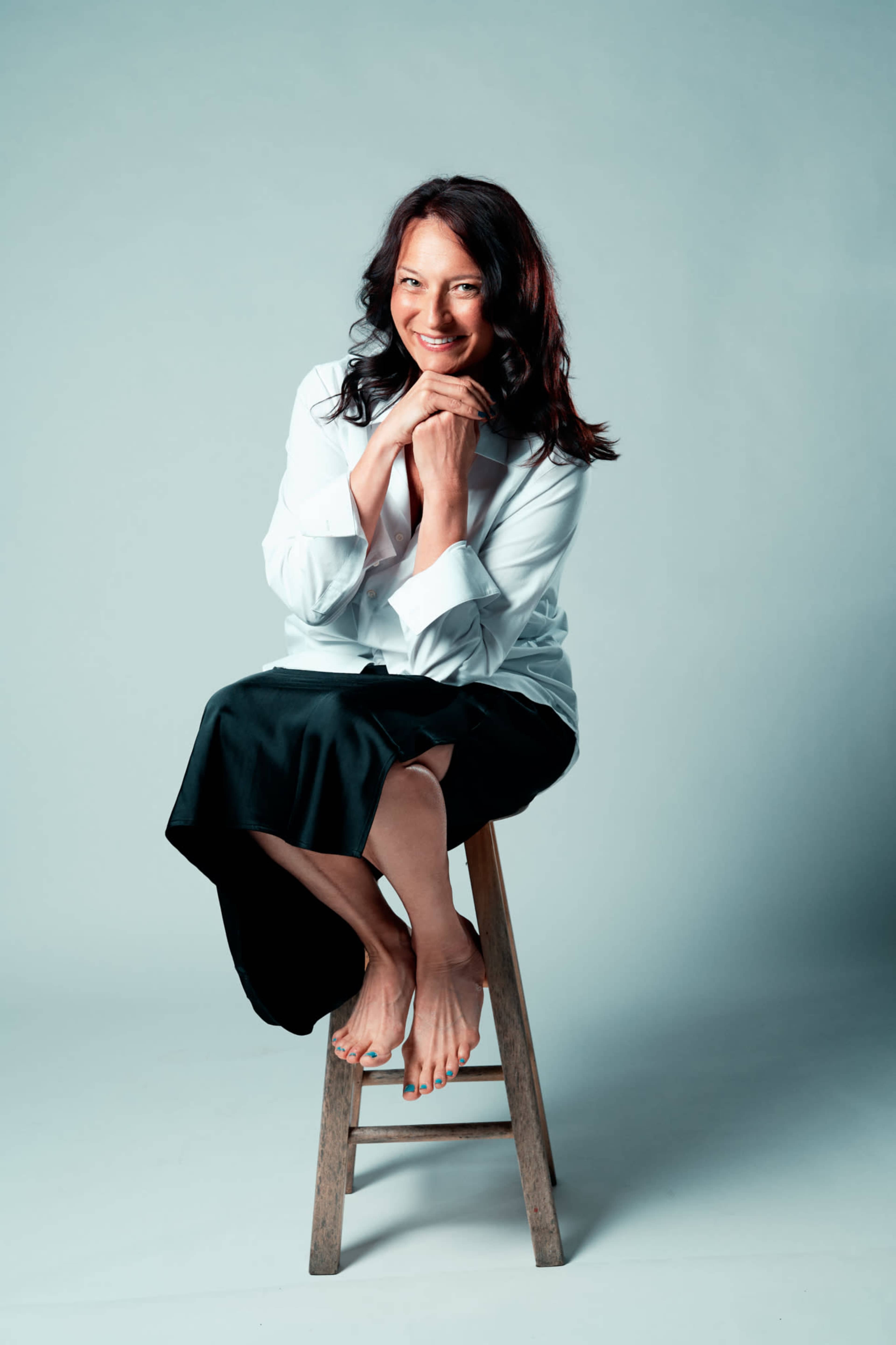 A woman sits on a wooden stool, smiling and resting her chin on her hands, wearing a white shirt and a black skirt.