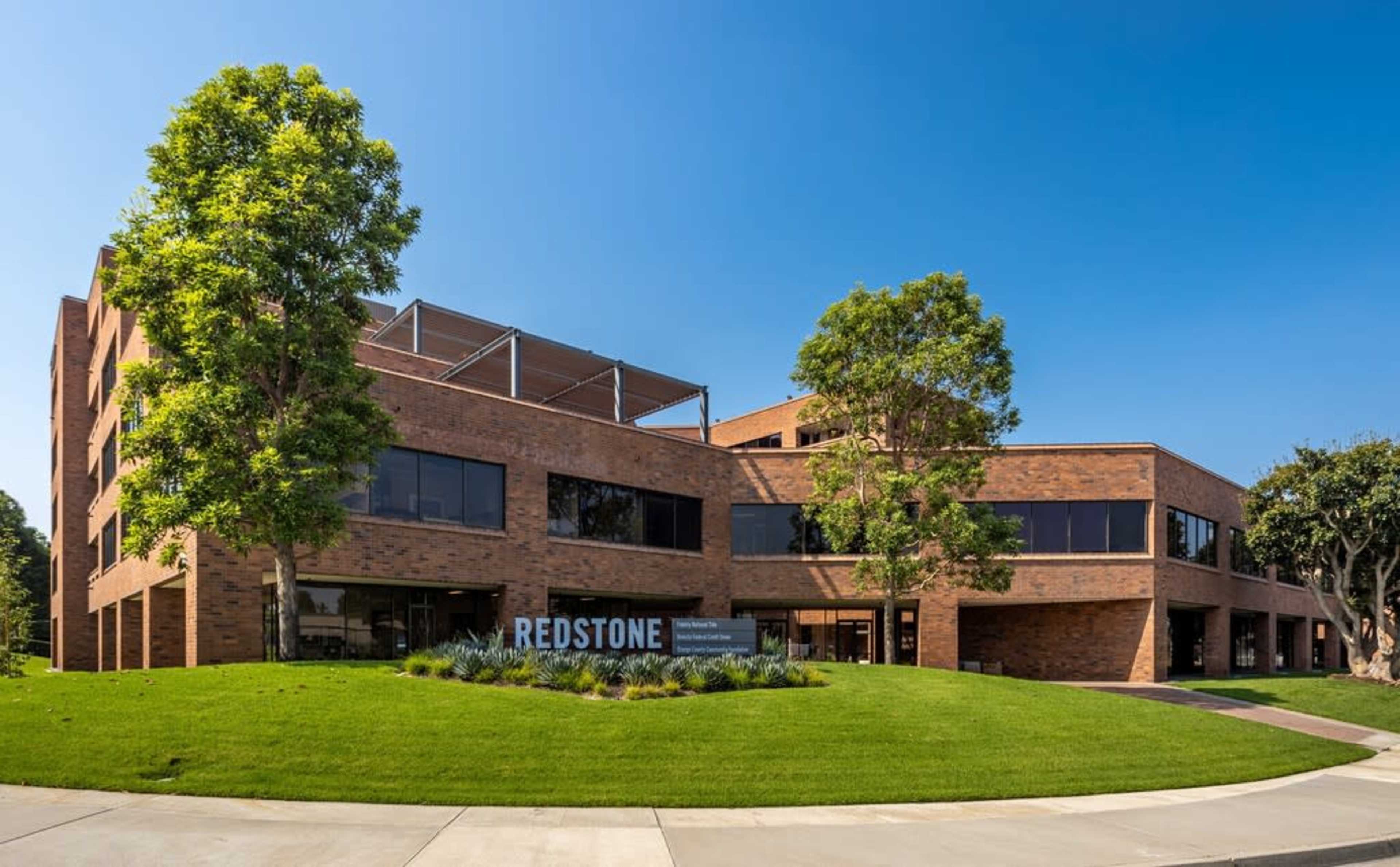 The image shows a modern brick building with large windows and a landscaped lawn, featuring a sign that reads "REDSTONE."