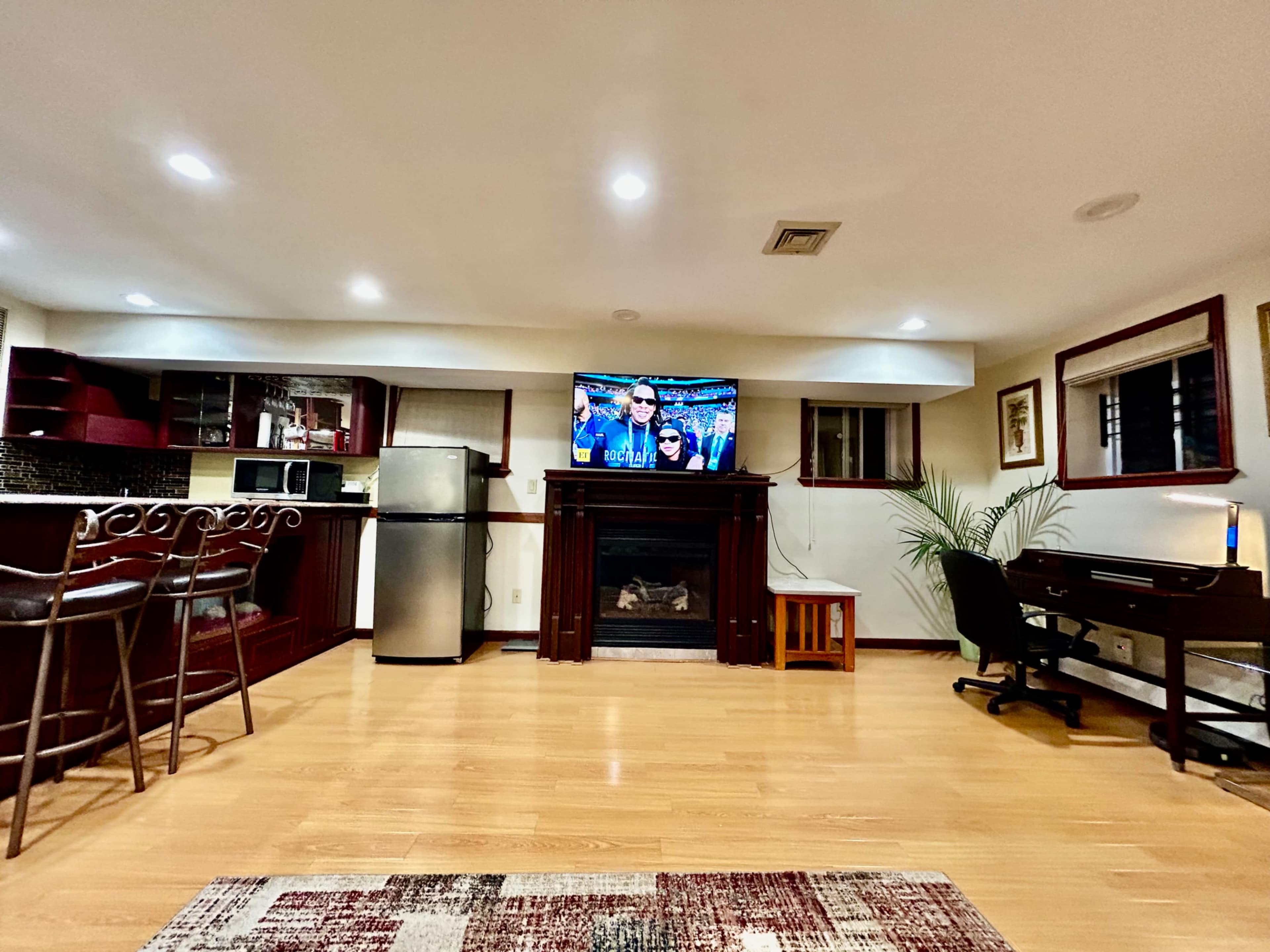 A basement room features a television above a fireplace, a bar area with stools, a desk, and a kitchenette.