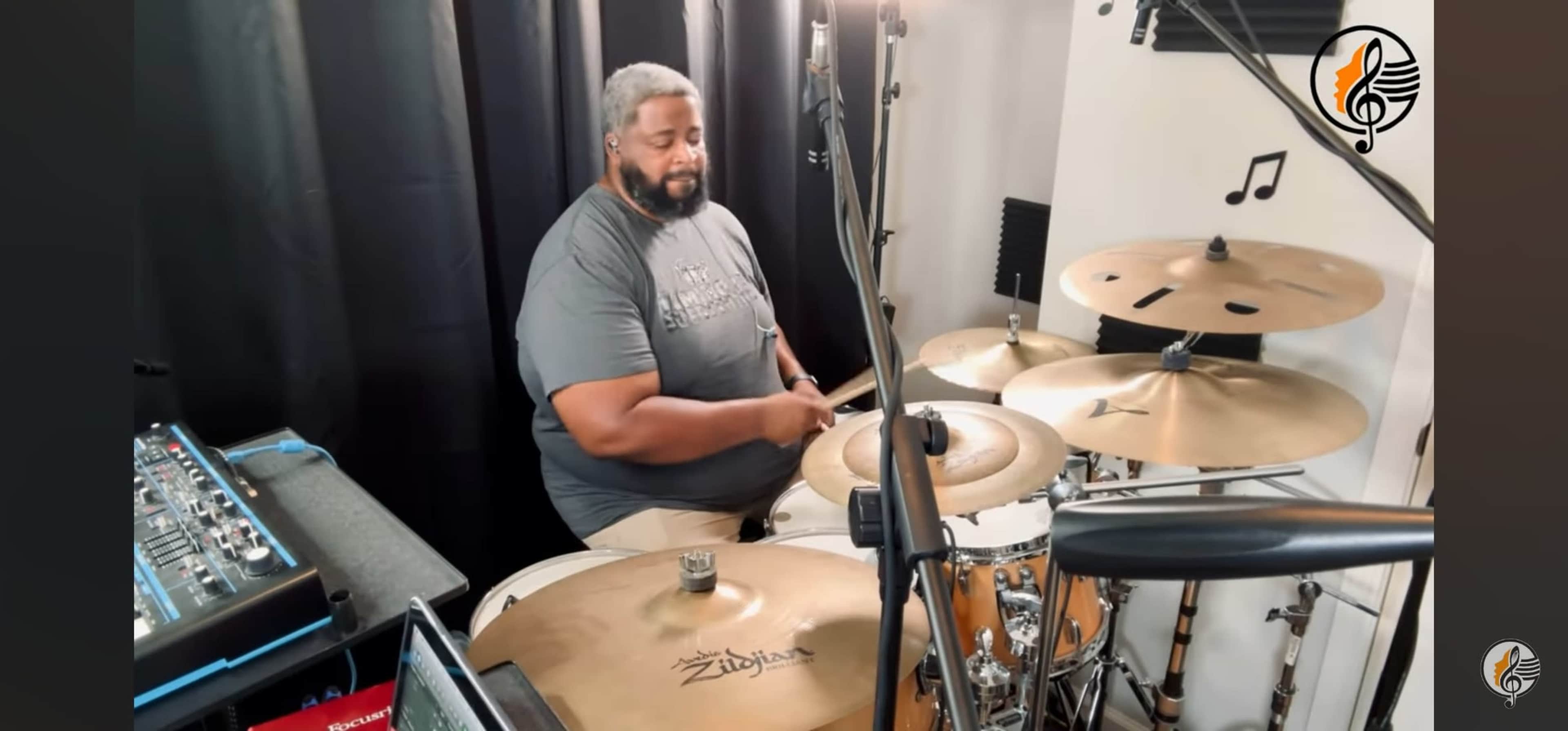 A man plays the drums in a studio setting with microphones and music equipment around him.