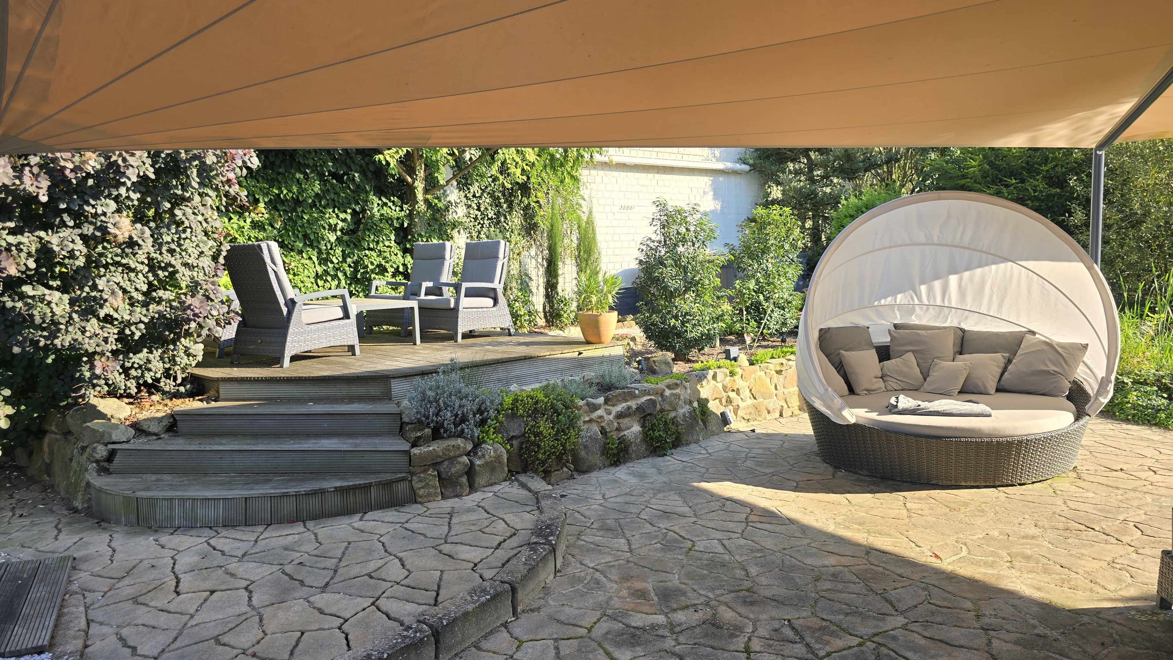 A shaded outdoor seating area features two chairs and a round lounge bed surrounded by greenery and stone pathways.