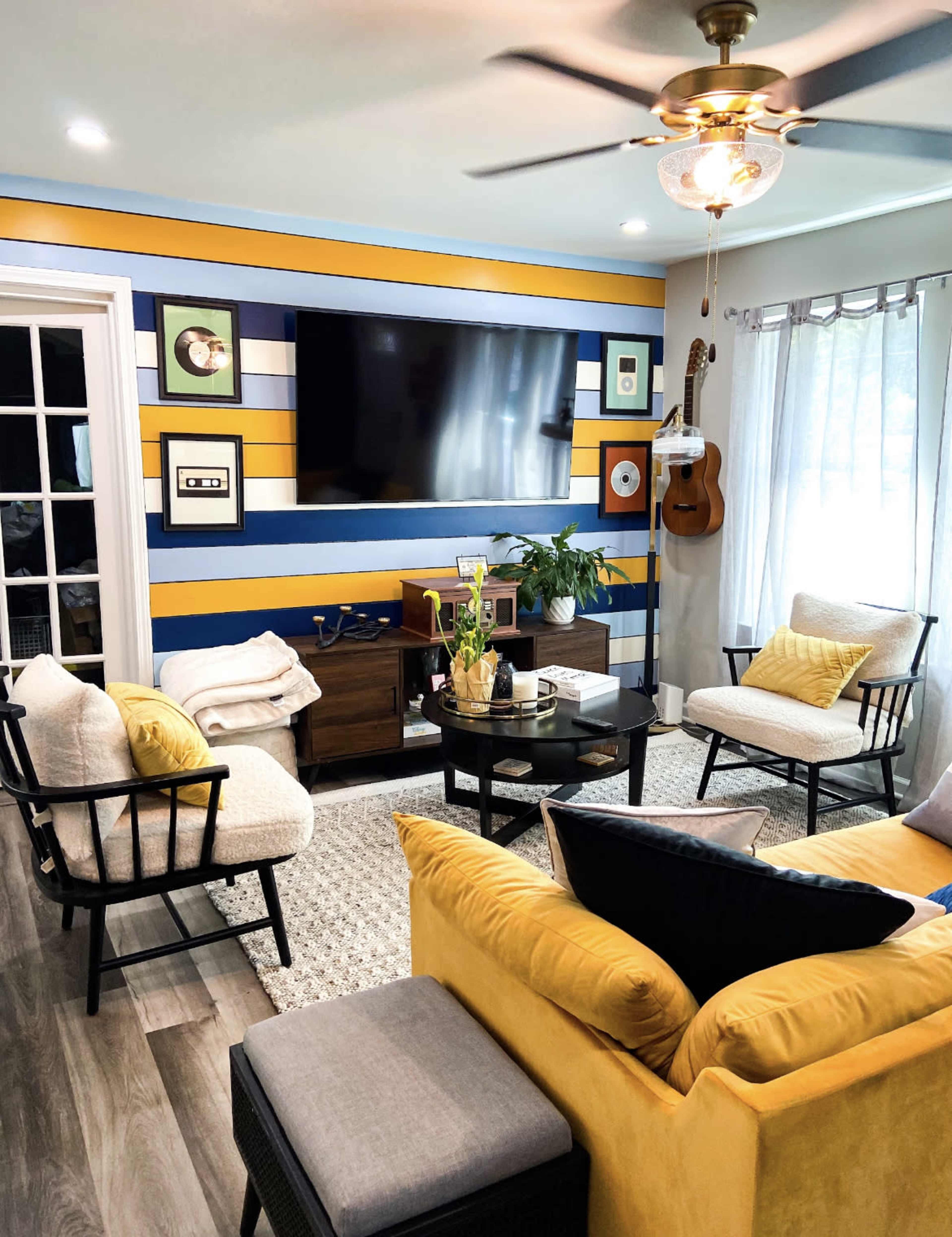 A brightly colored living room features a striped accent wall, a large flat-screen TV, comfortable seating, and various decorative elements including plants and a guitar.