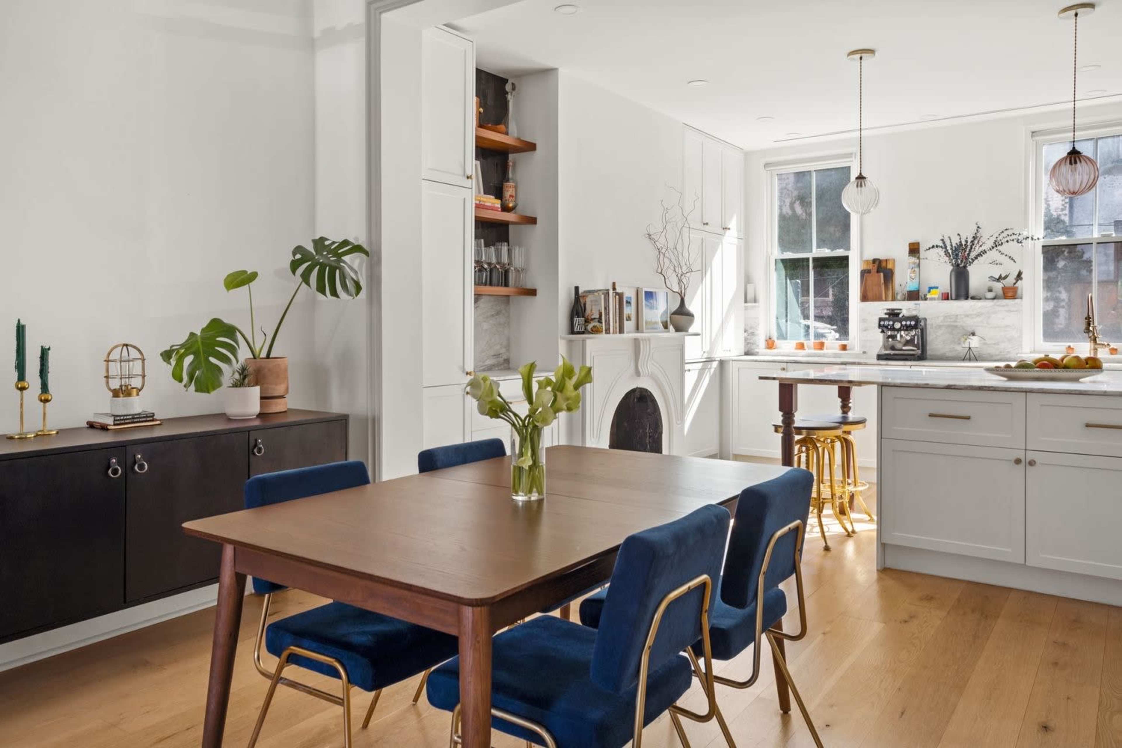 The image shows a modern dining area with a wooden table surrounded by blue chairs, a dark sideboard, and a kitchen visible in the background with white cabinetry and a marble countertop.