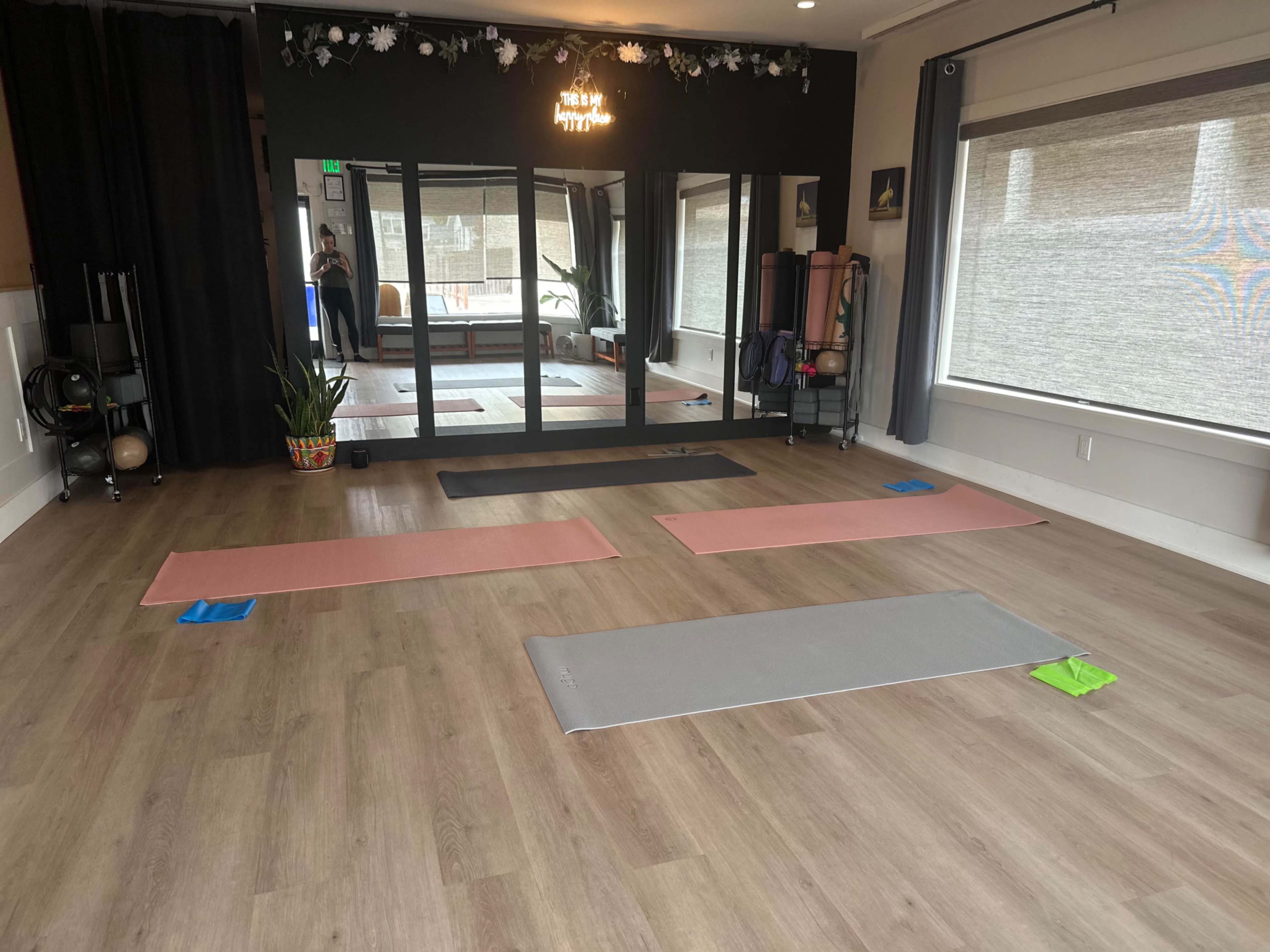 The image shows a yoga or fitness studio with several exercise mats arranged on a wooden floor, positioned near mirrors and various workout equipment.