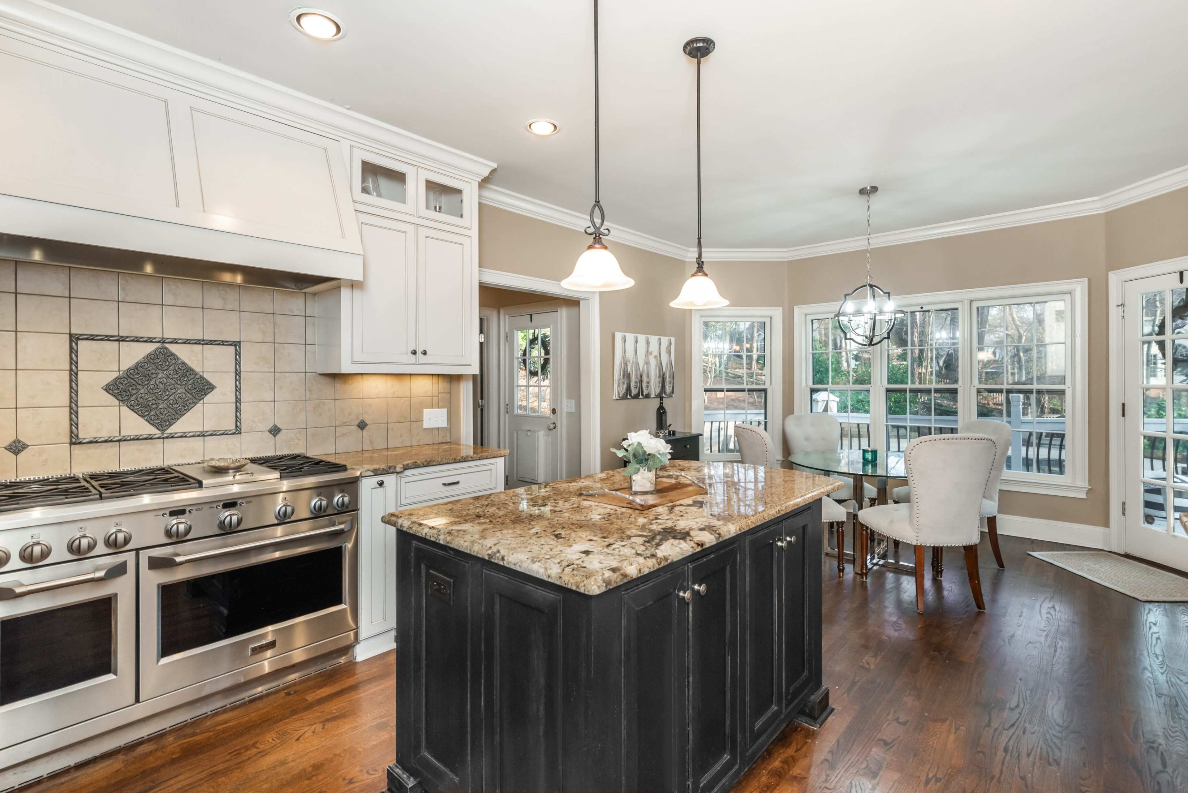 A spacious kitchen features a central island with a dark finish, granite countertops, and stainless steel appliances, surrounded by large windows and a dining area.