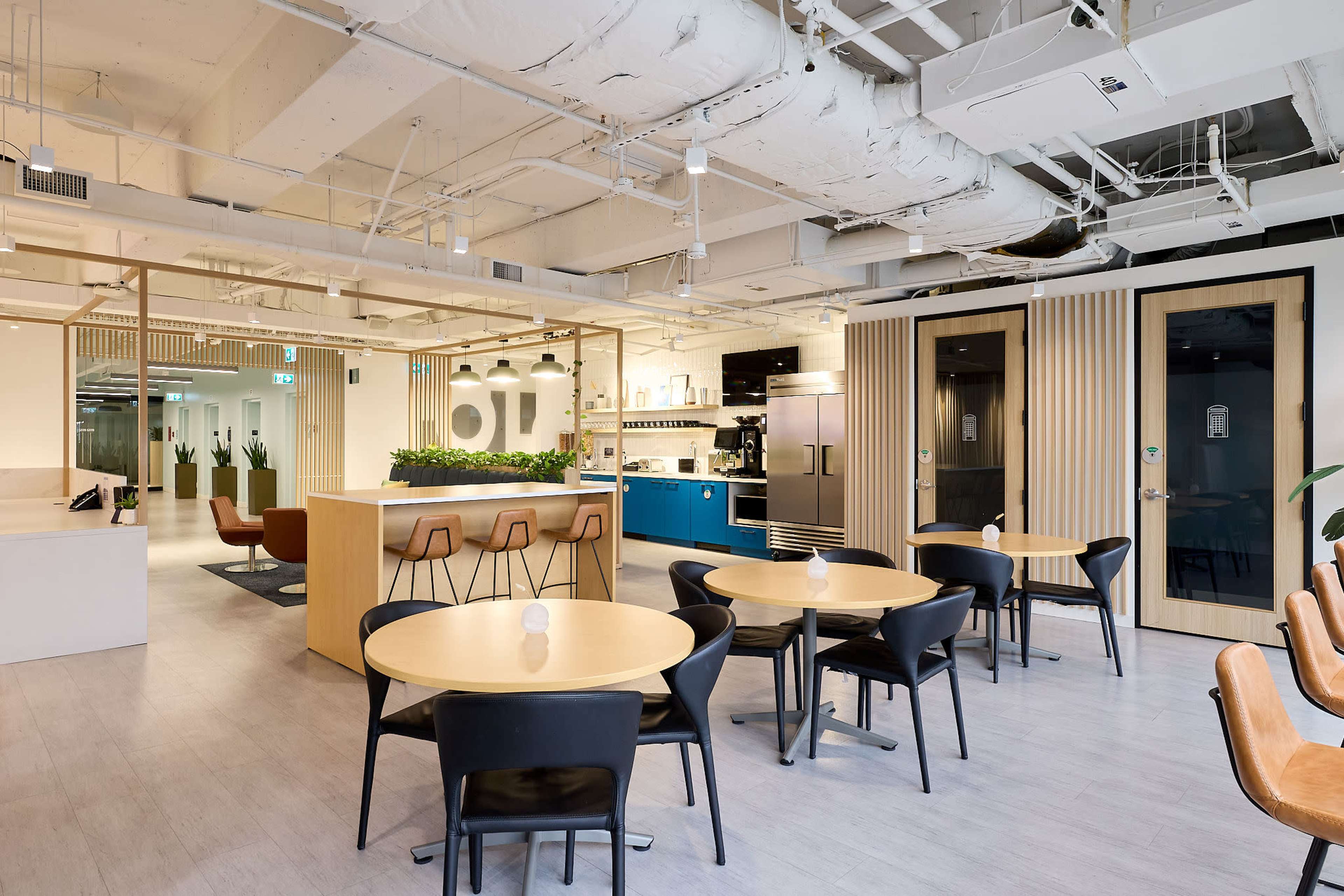 The image shows a modern office space featuring a kitchen area, multiple round tables with black chairs, and a few plants along the walls.