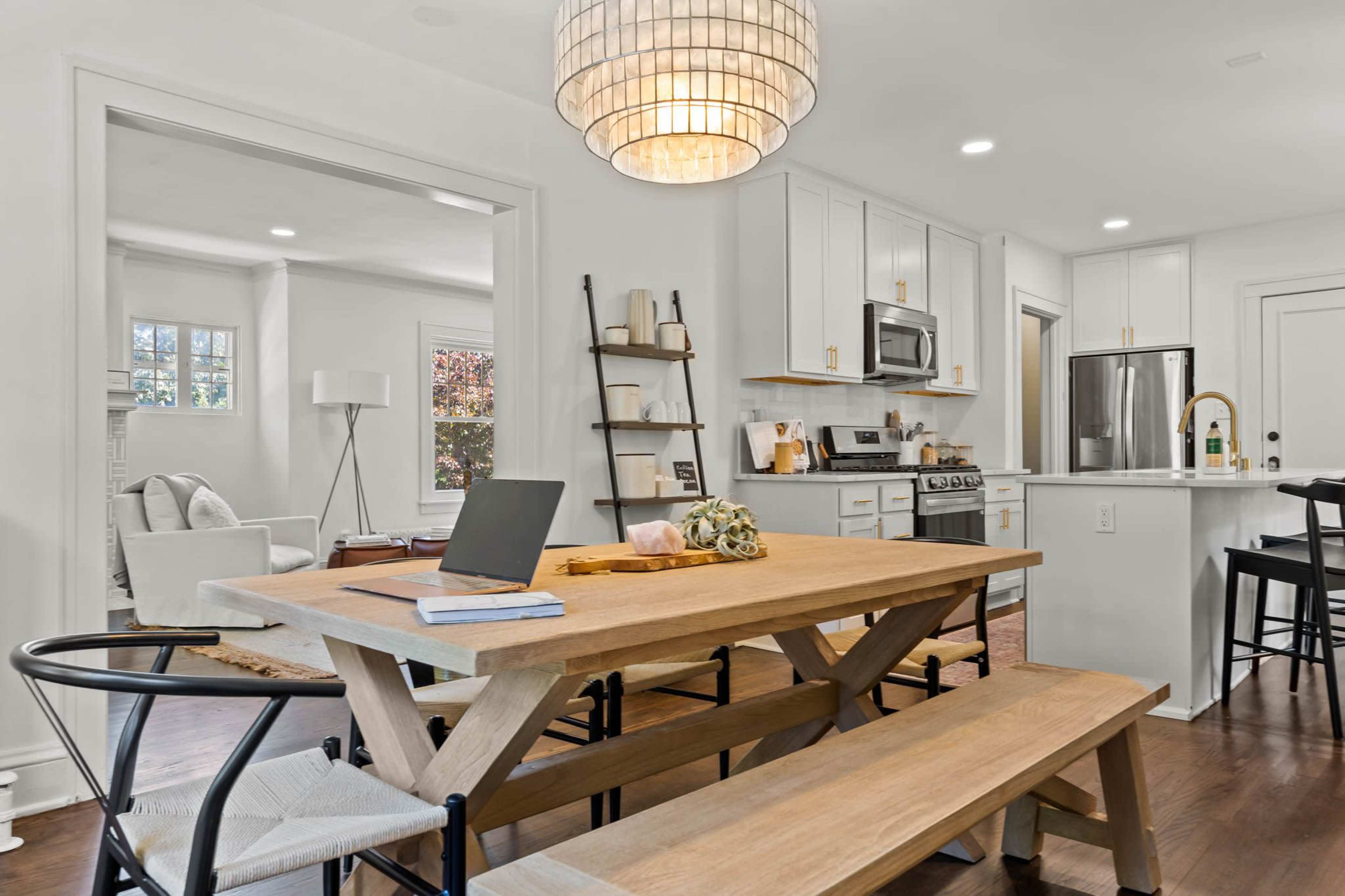 A modern kitchen and dining area featuring a wooden table with benches, a laptop, and a chandelier hanging above.