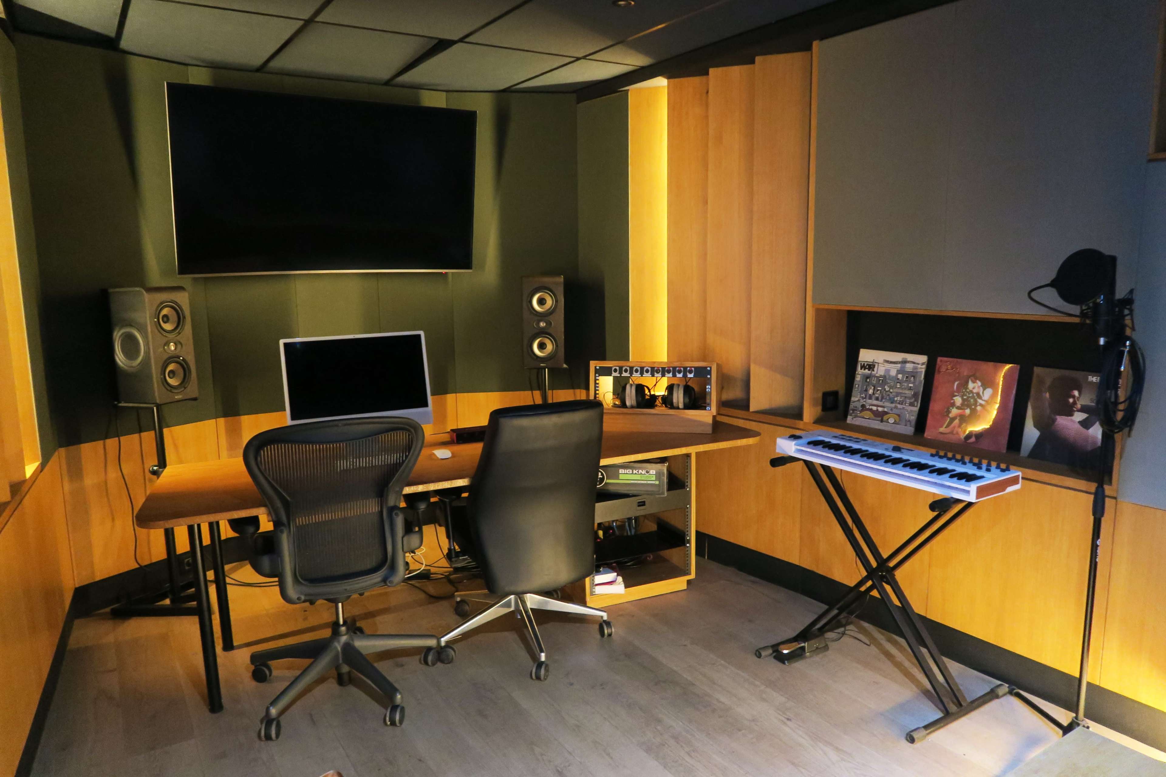 The image shows a music studio with a large desk, computer, speakers, and a keyboard, featuring wooden paneling and dim lighting.