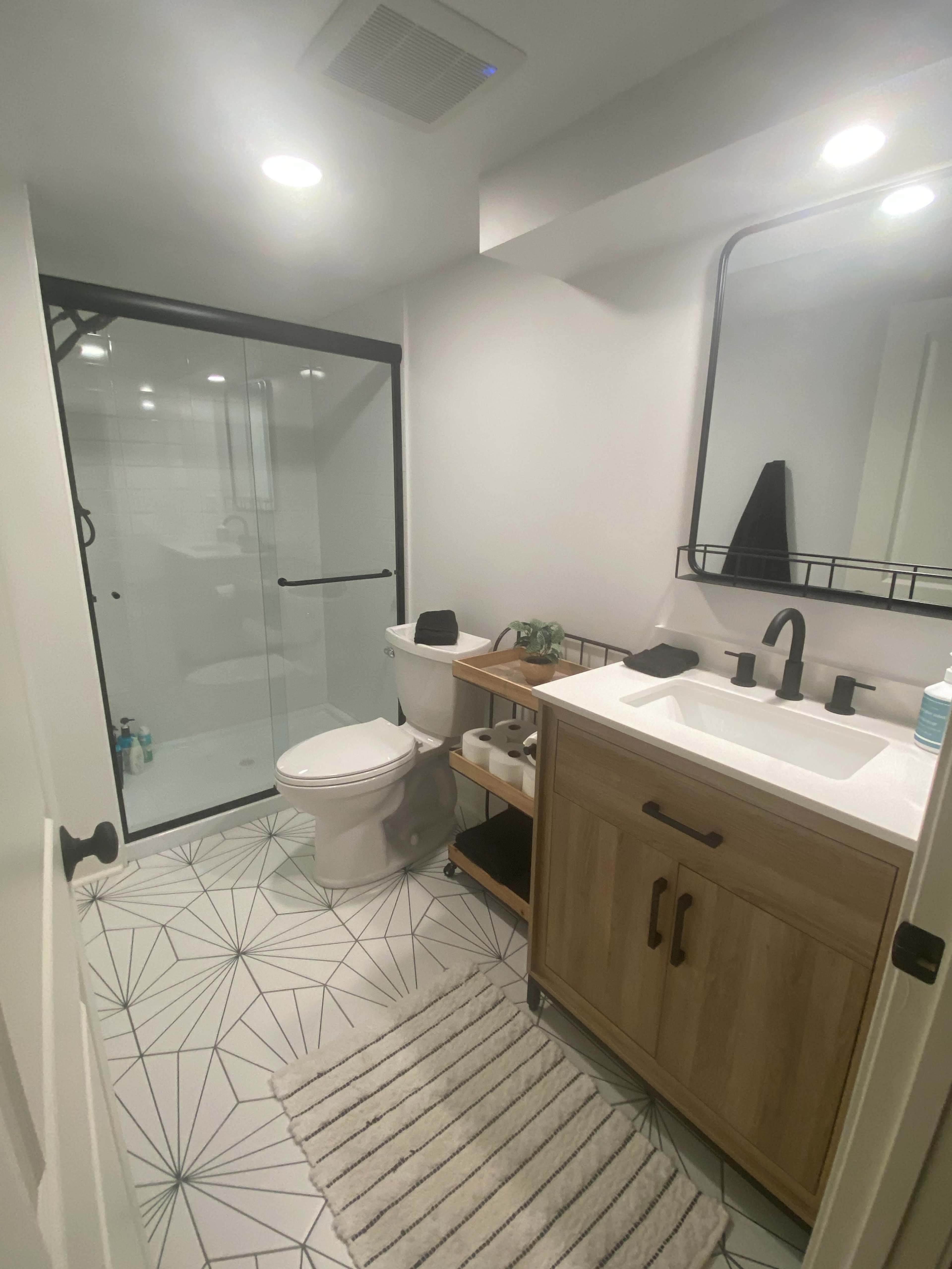 The image shows a modern bathroom featuring a glass shower enclosure, a wooden vanity with a sink, a toilet, and geometric patterned floor tiles.
