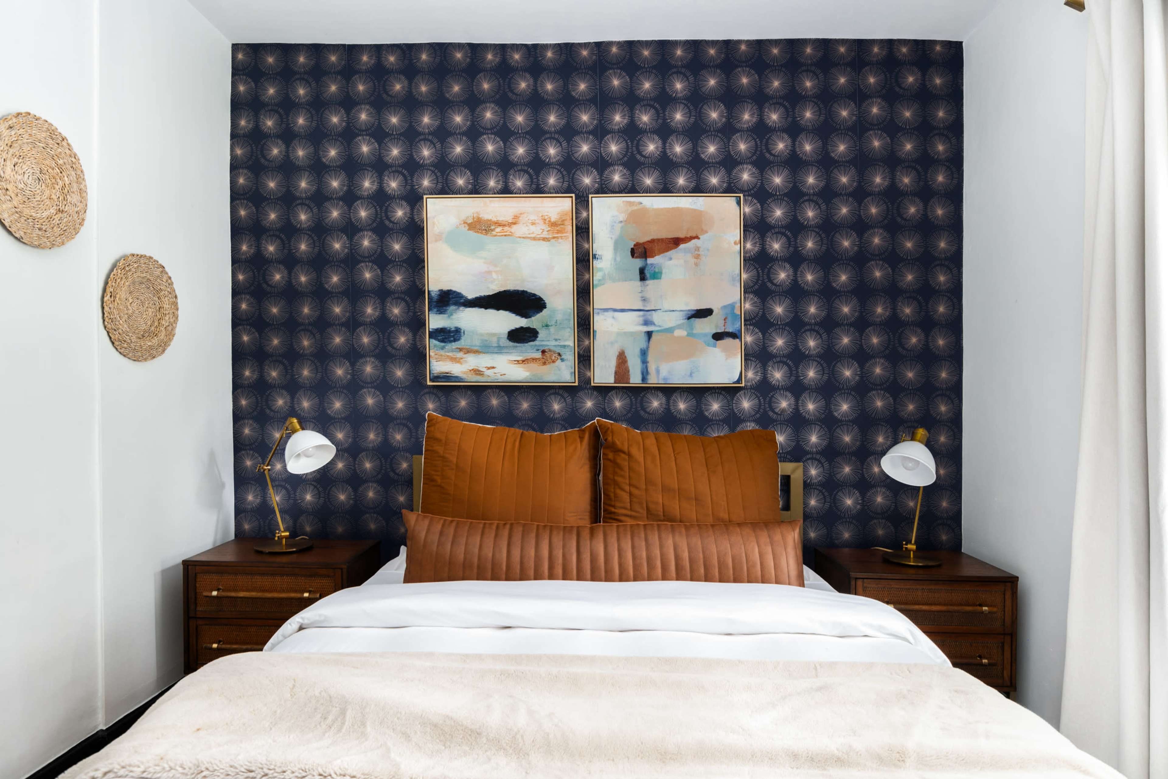 The image displays a modern bedroom featuring a bed with brown pillows, two framed artworks on a patterned blue wall, and bedside tables with table lamps.