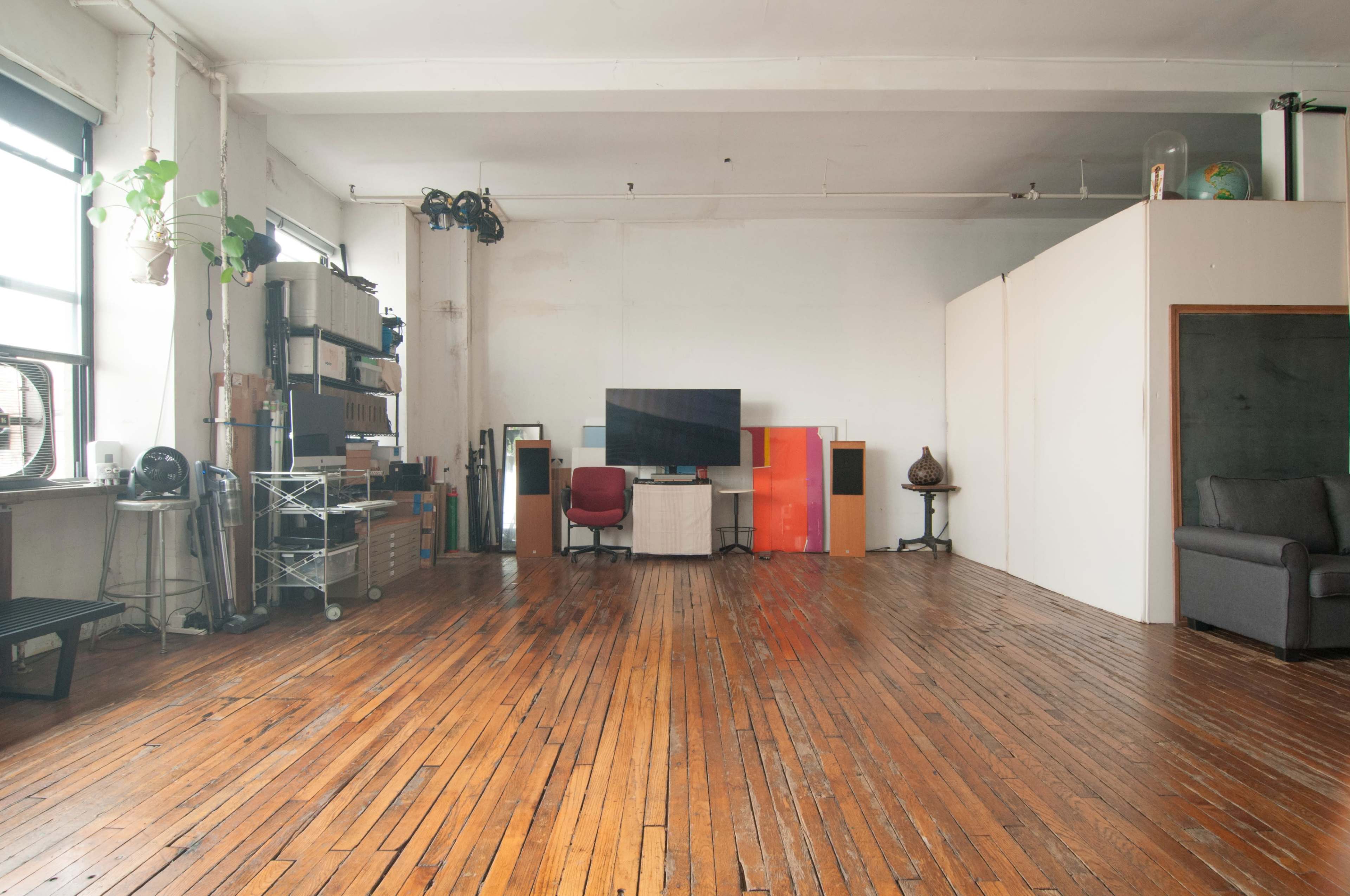The image shows a spacious, minimally furnished room with hardwood floors, a television, and various storage items along the walls.