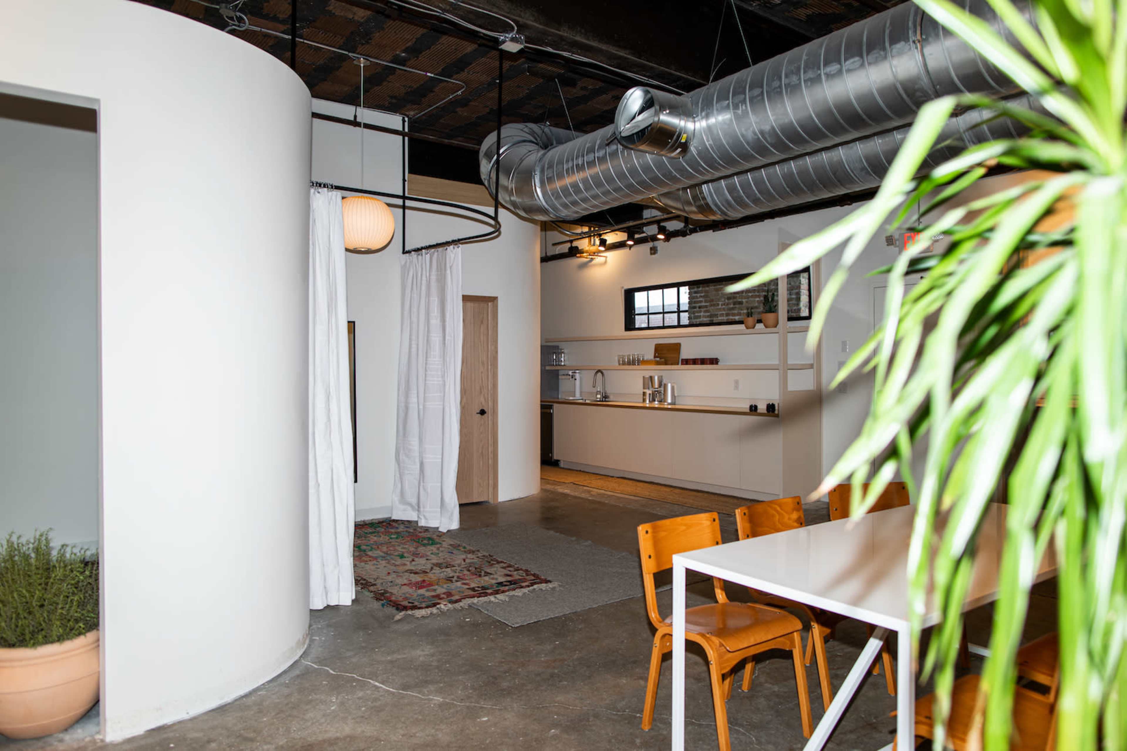 The image shows a modern interior space featuring a circular partition, a dining table with wooden chairs, a kitchenette in the background, and potted plants.