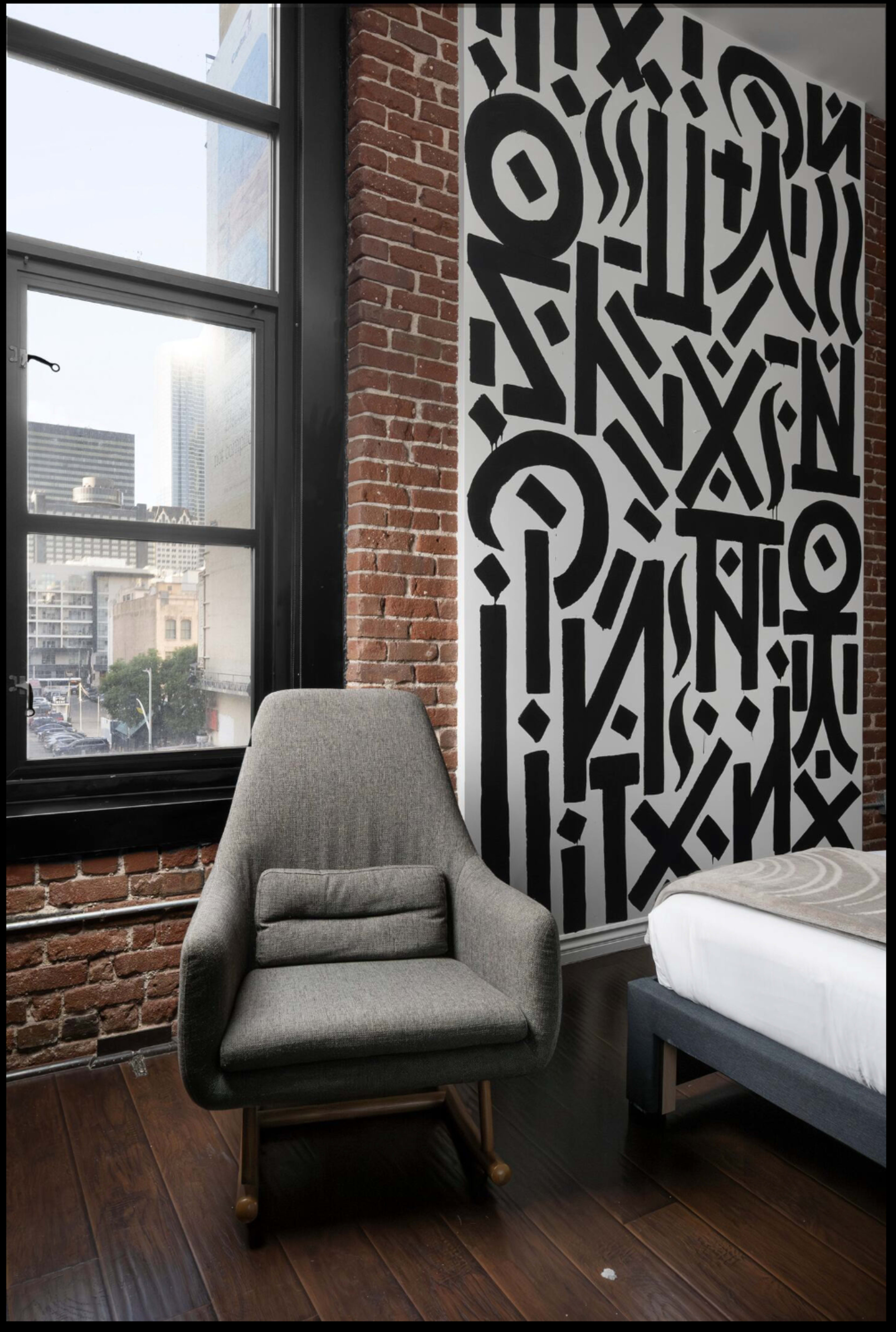 A modern gray chair sits beside a window in a room featuring a brick wall and a large, black graphic mural.