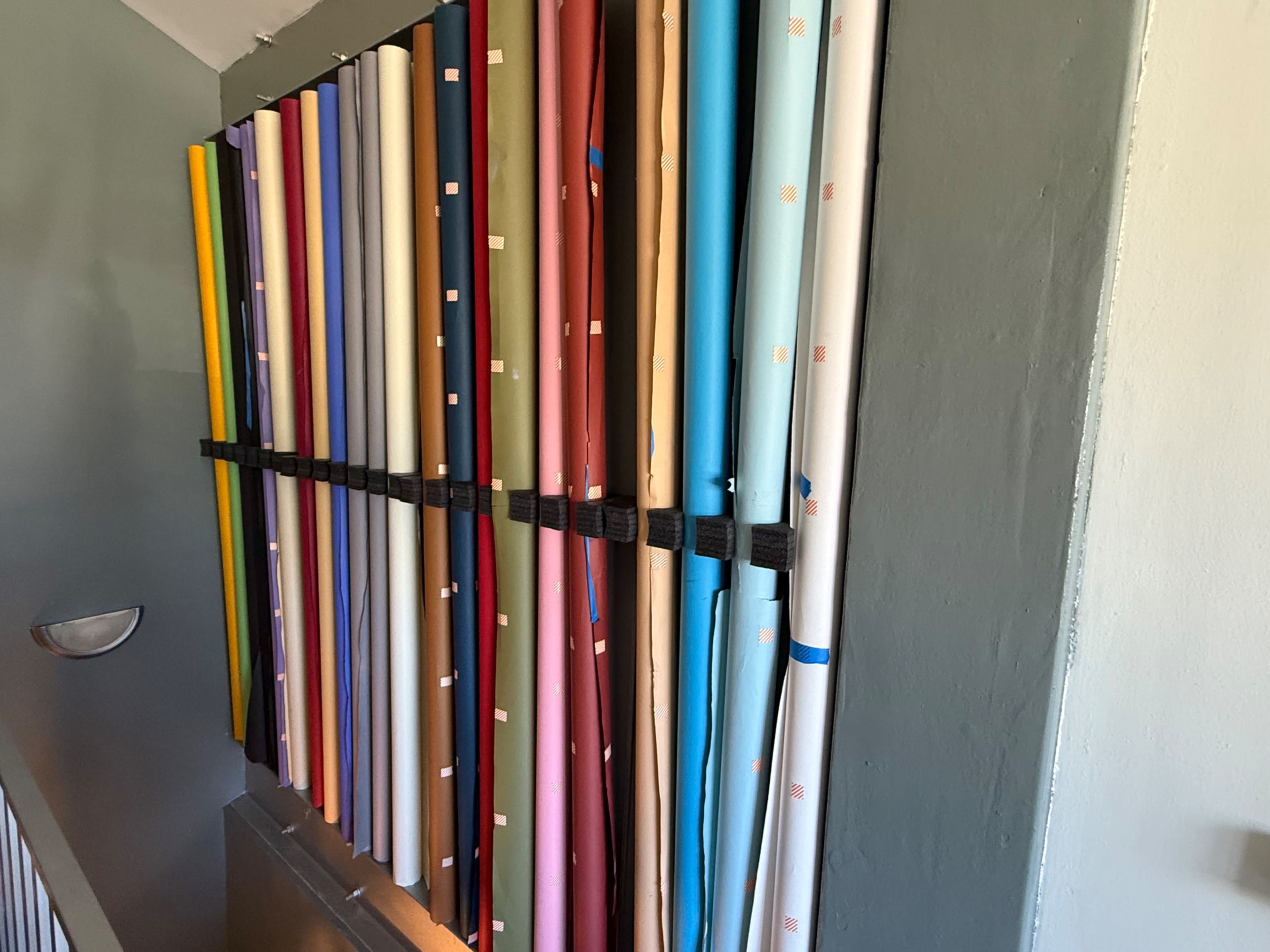The image shows a vertical arrangement of rolled paper or fabric in various colors secured by straps against a wall.
